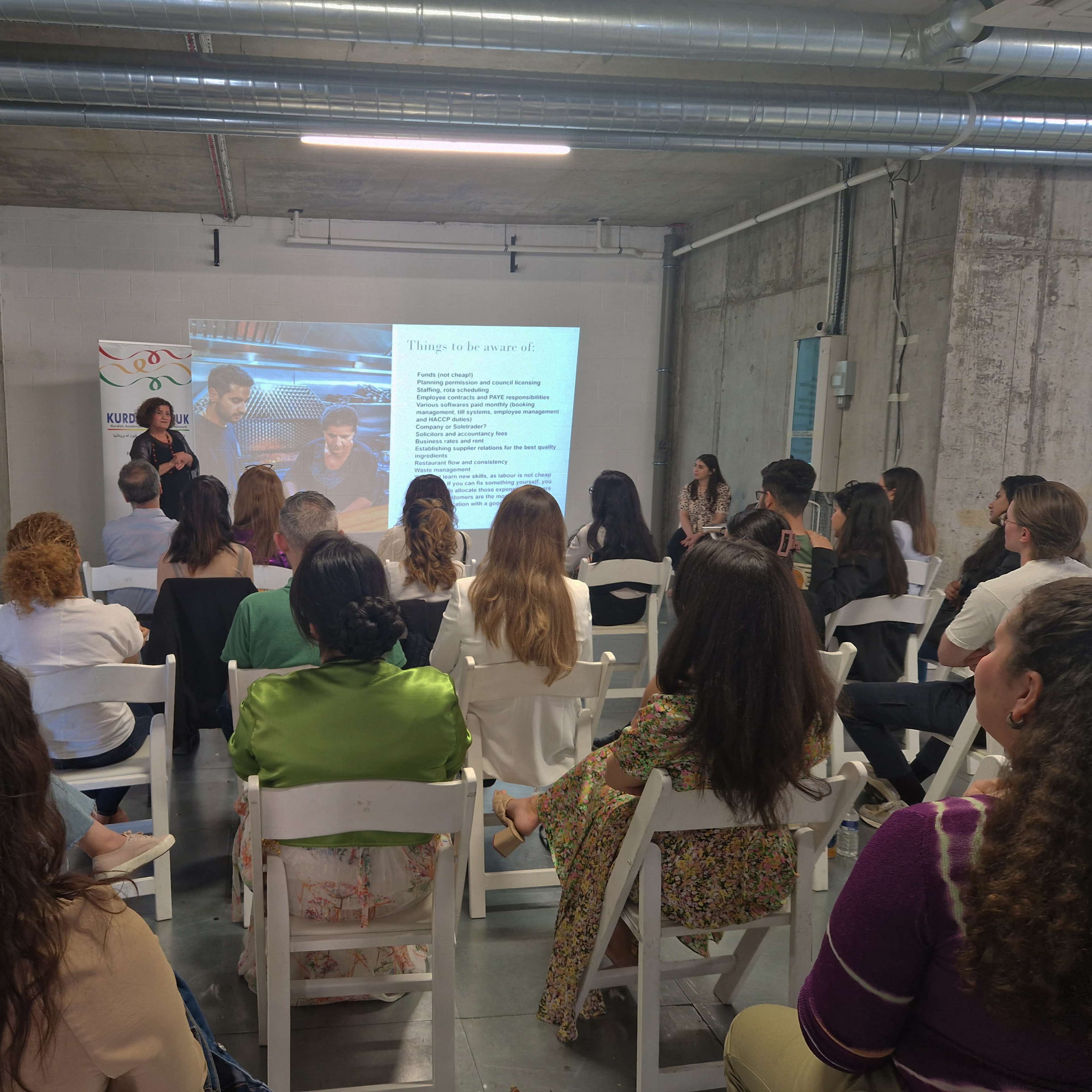 A speaker presents information to an audience seated in a conference room with a projector display in the background.