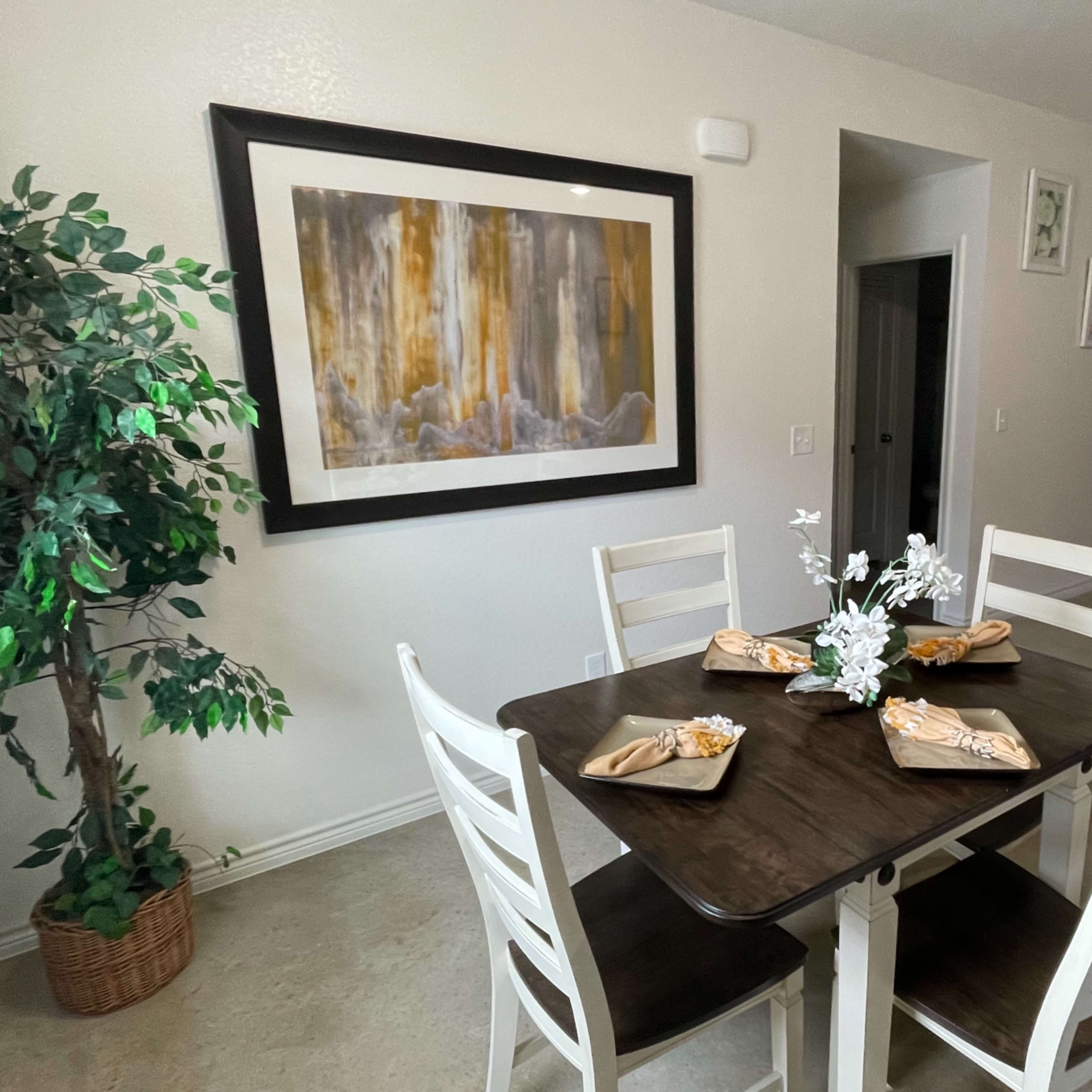 A dining table set with decorative napkins and flowers is positioned near a framed artwork in a kitchen area.