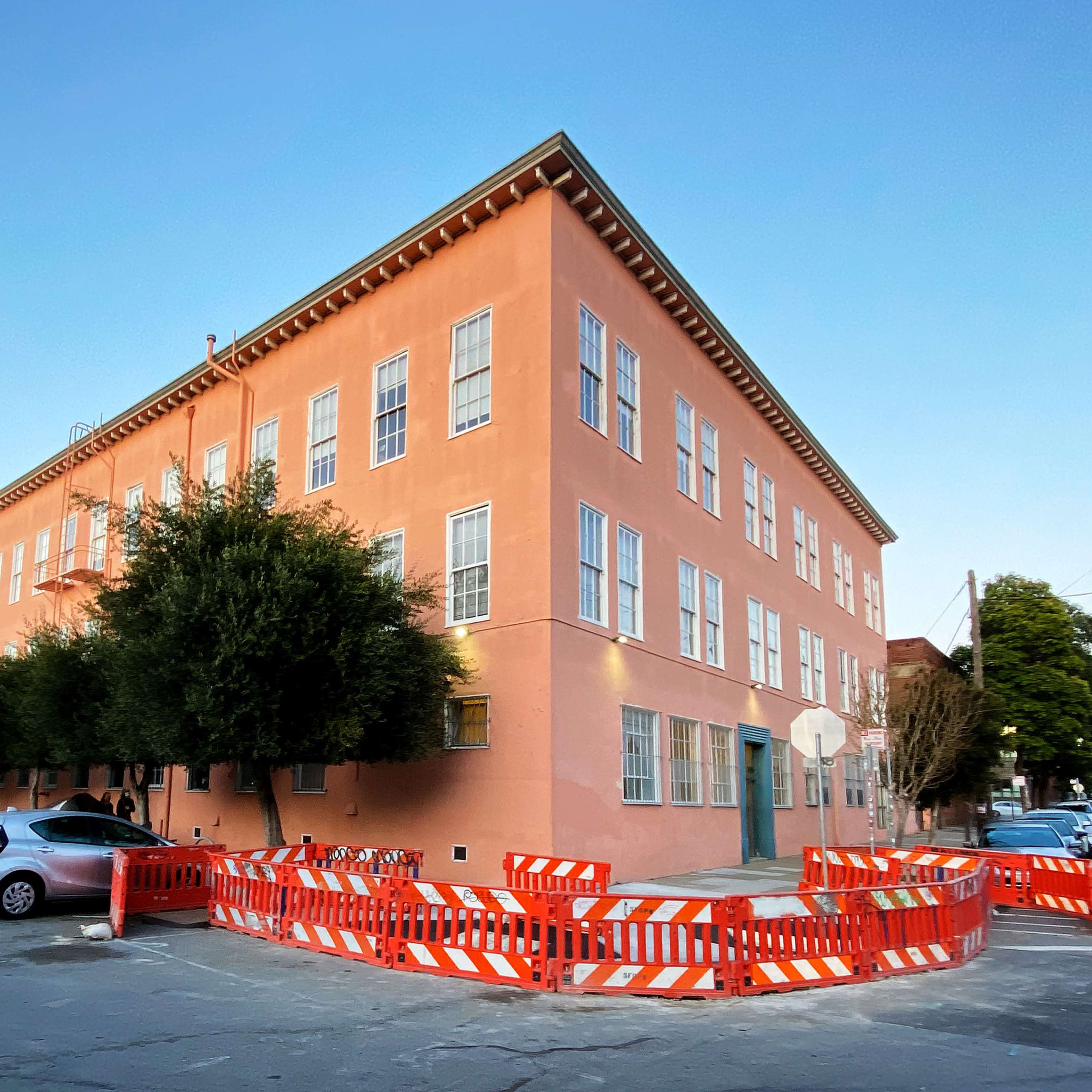 A pink building with large windows is located at the corner of a street, surrounded by orange construction barriers and several parked cars.