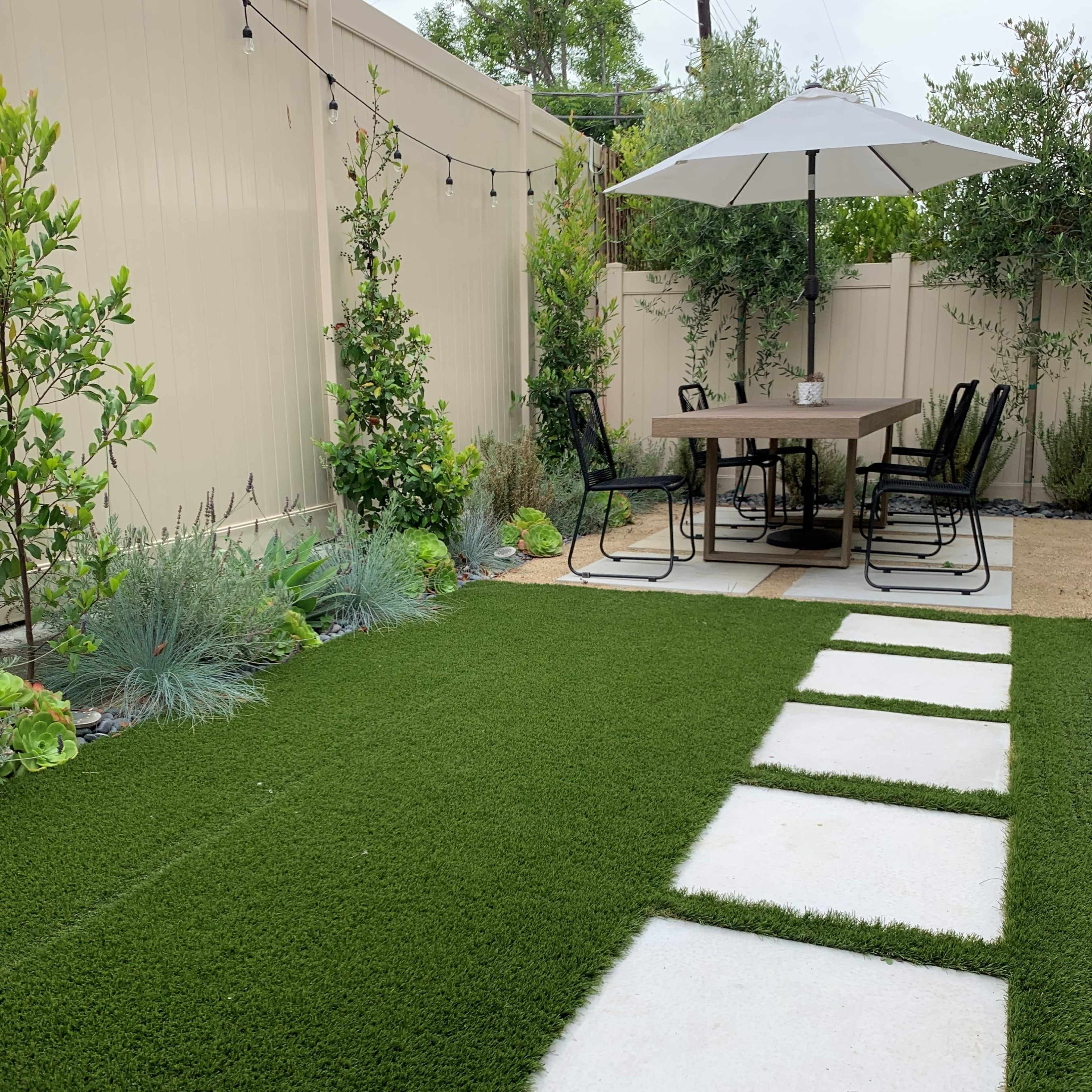 A patio area features a dining table with chairs under a large umbrella, surrounded by greenery and a path of stone pavers on artificial turf.