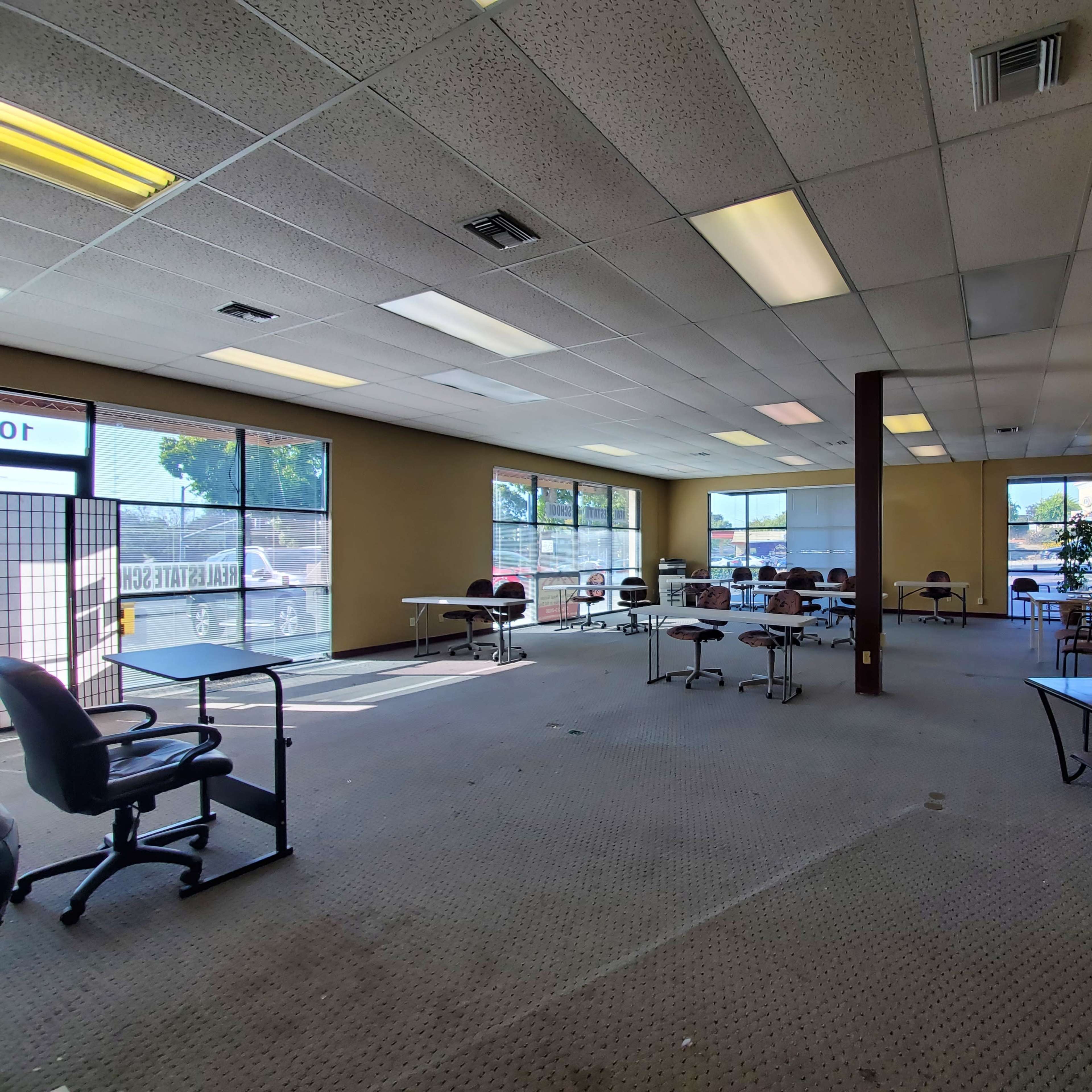 An empty commercial space with several tables and chairs arranged throughout, large windows letting in natural light, and a view of a parking lot outside.
