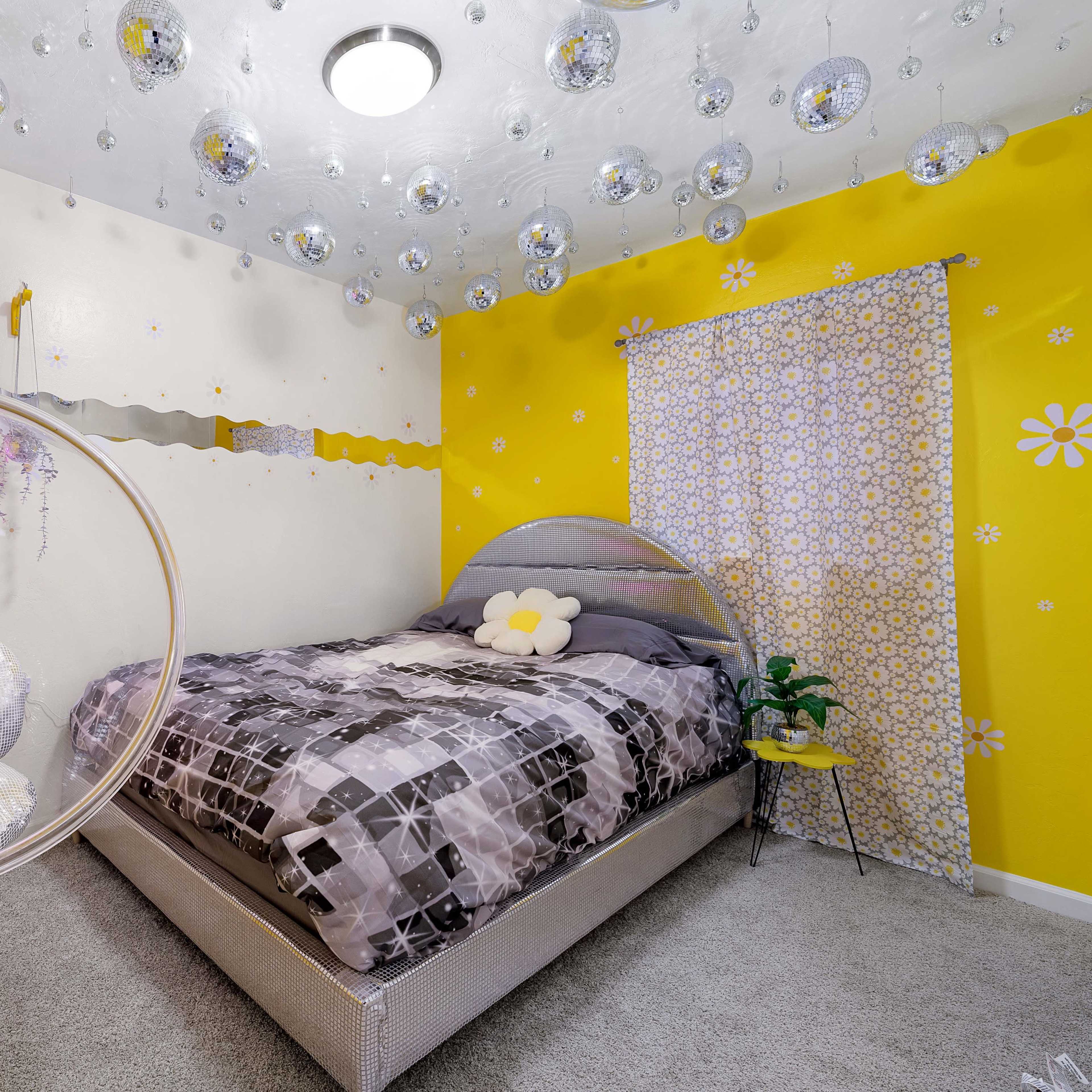 A bedroom features yellow walls adorned with flower decals, a large bed with a patterned comforter, and a hanging circular sign that reads "DISCO MADE ME DO IT," surrounded by mirrored disco balls.