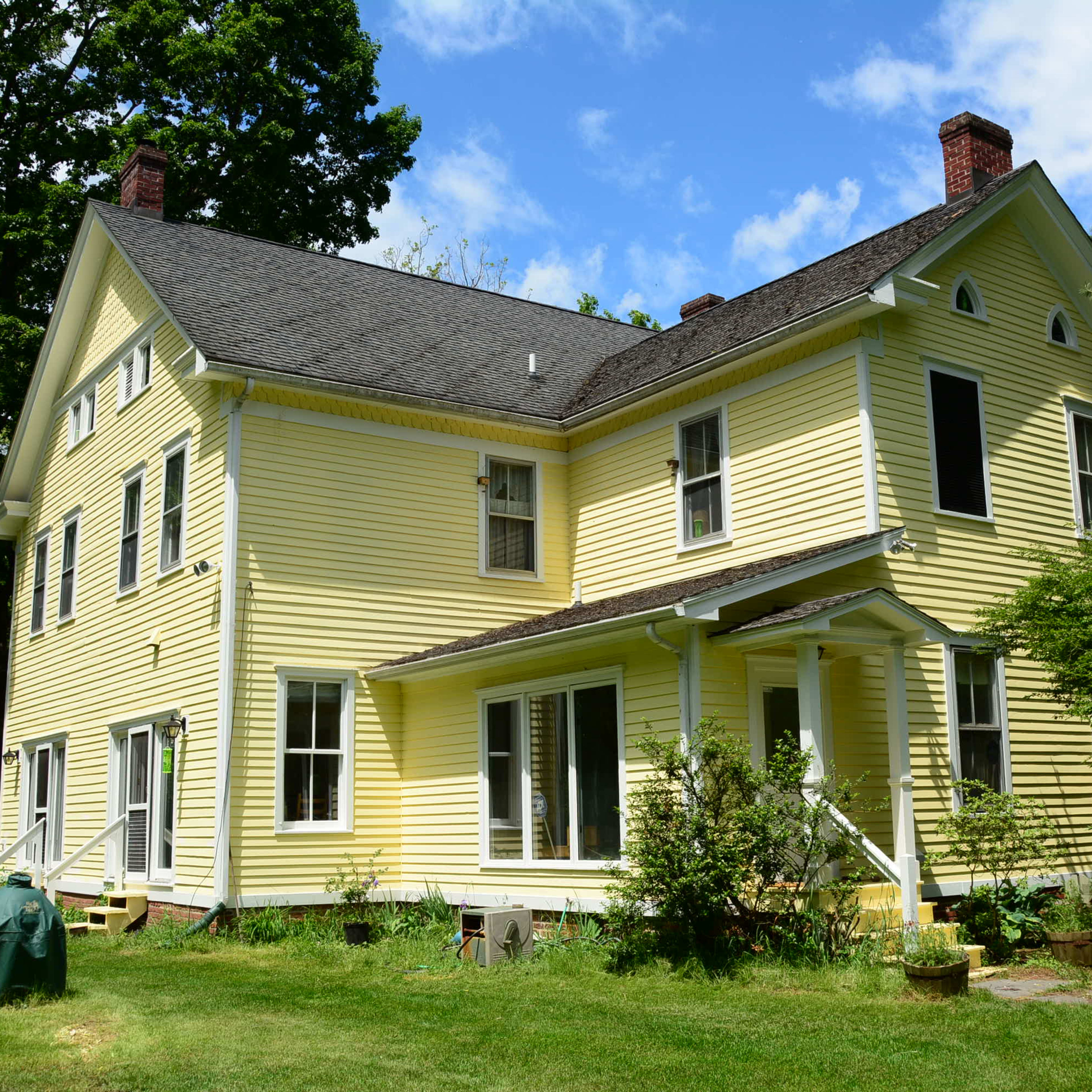 A two-story yellow house with multiple windows and a steep roof is surrounded by green grass and trees.