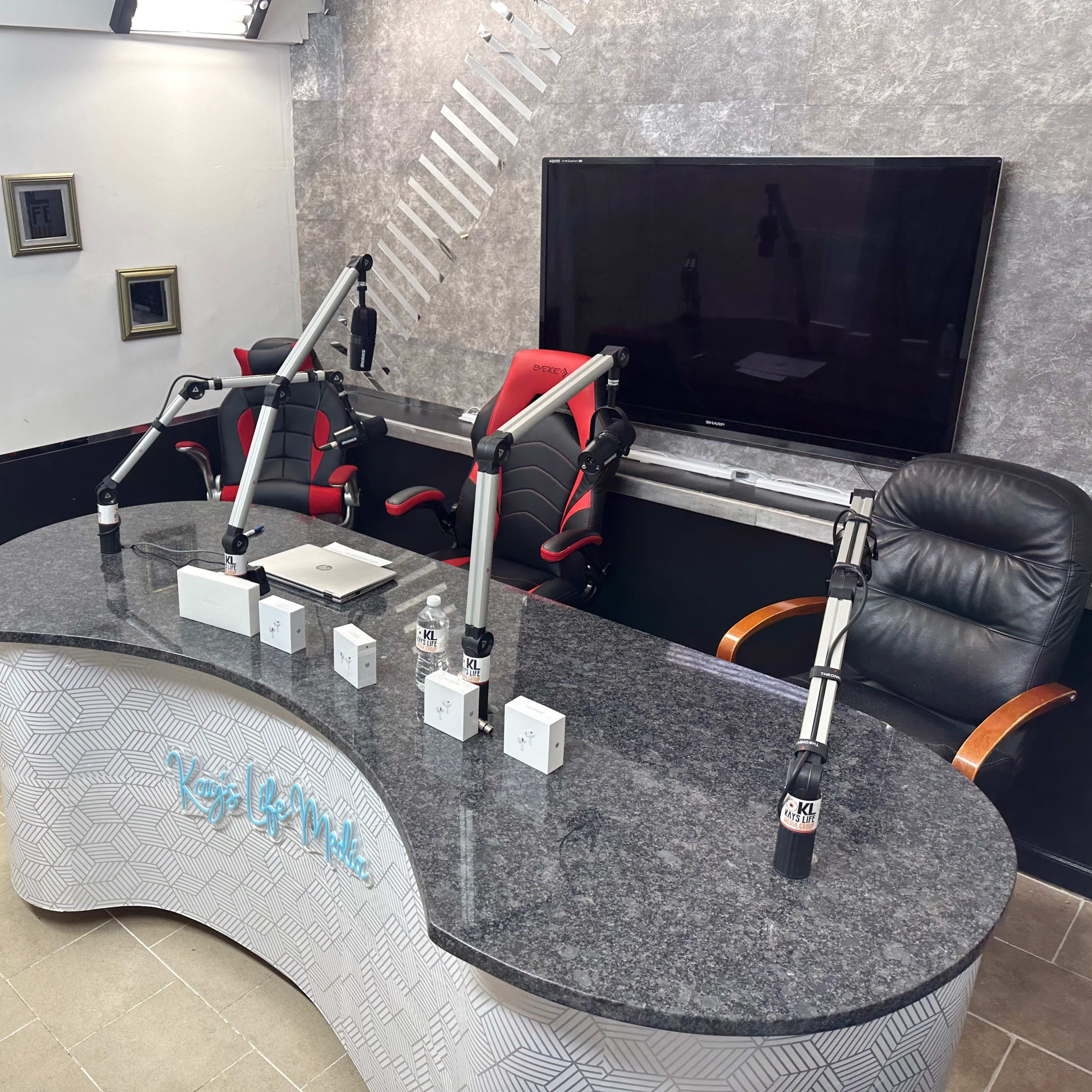 A modern recording studio with a curved granite table, three microphone stands, a laptop, and a large television screen on the wall.