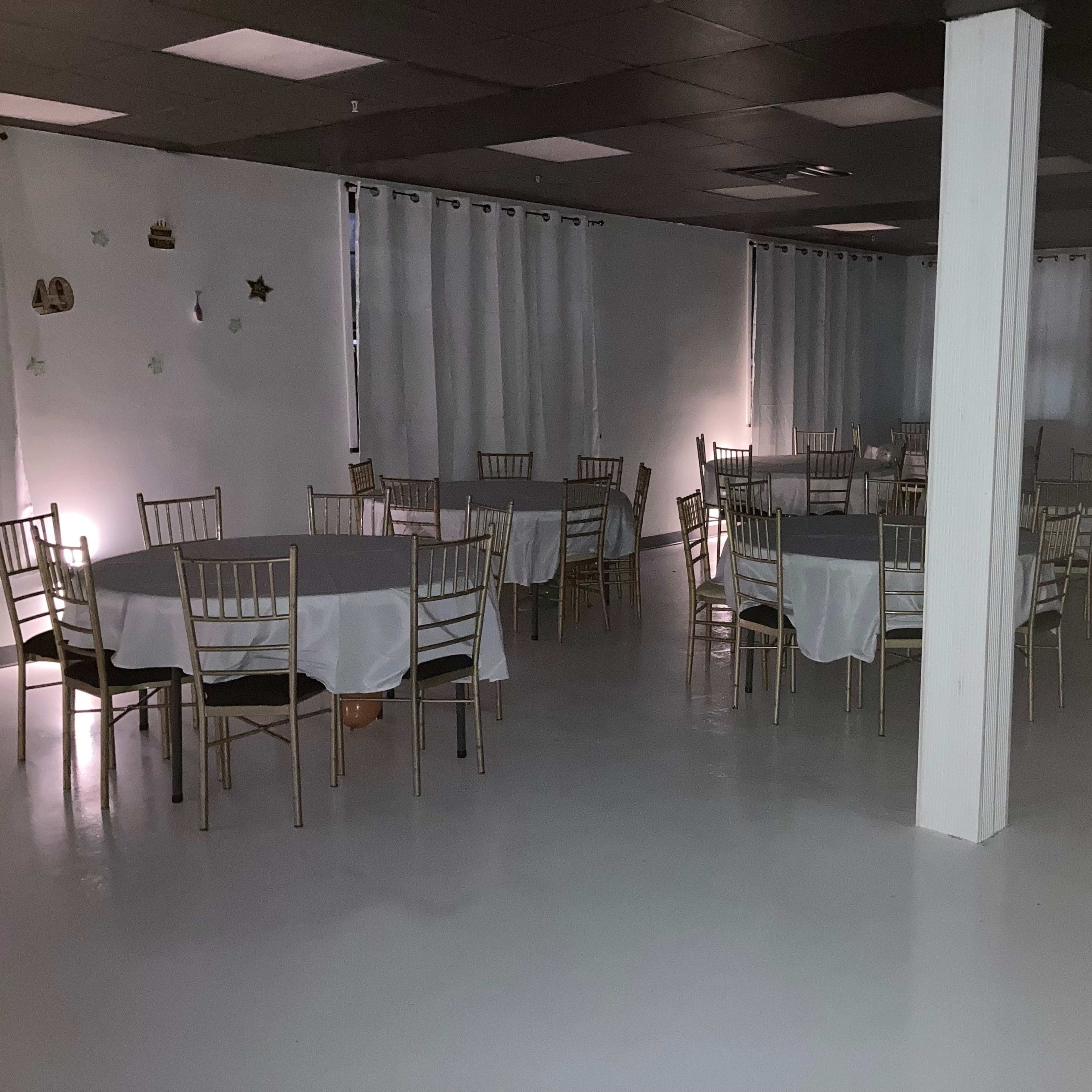 A dimly lit hall with several round tables covered with white tablecloths and gold chairs arranged throughout the space.