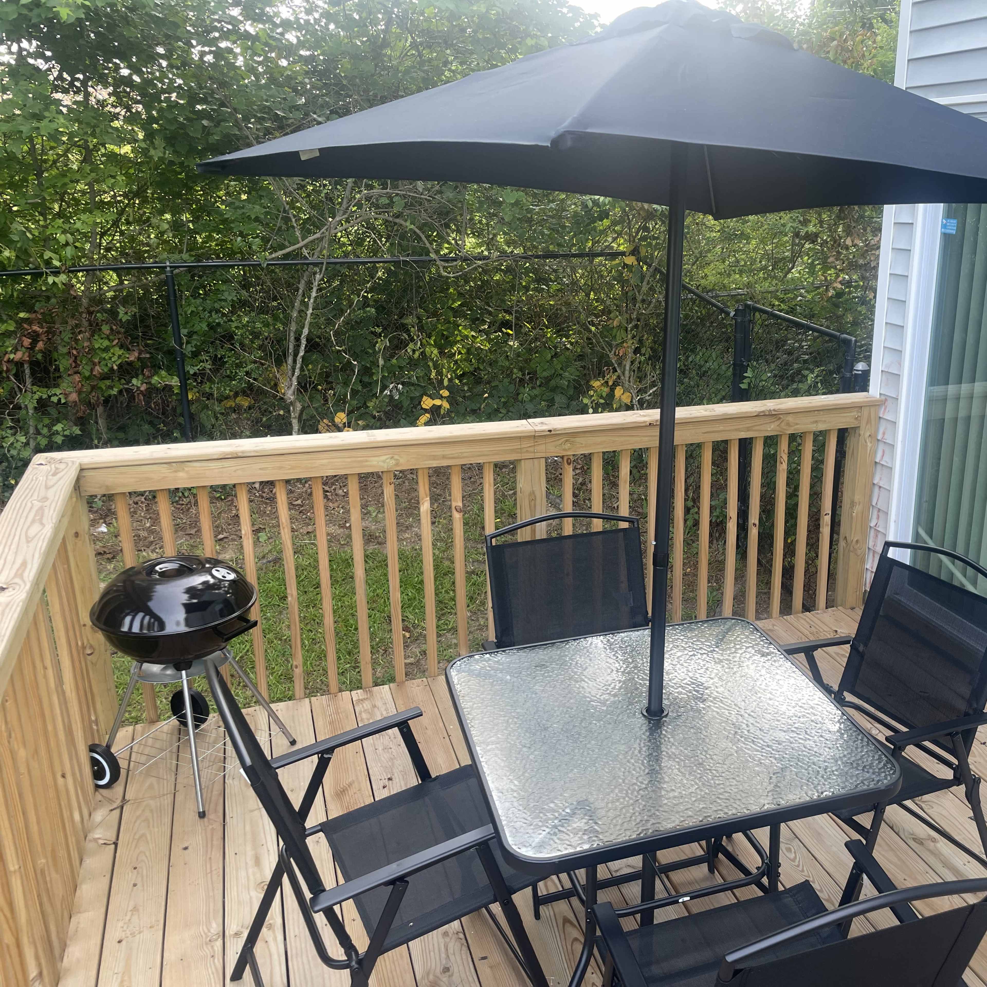 A black patio umbrella shades a square glass table surrounded by six black chairs on a wooden deck, with a small grill nearby.