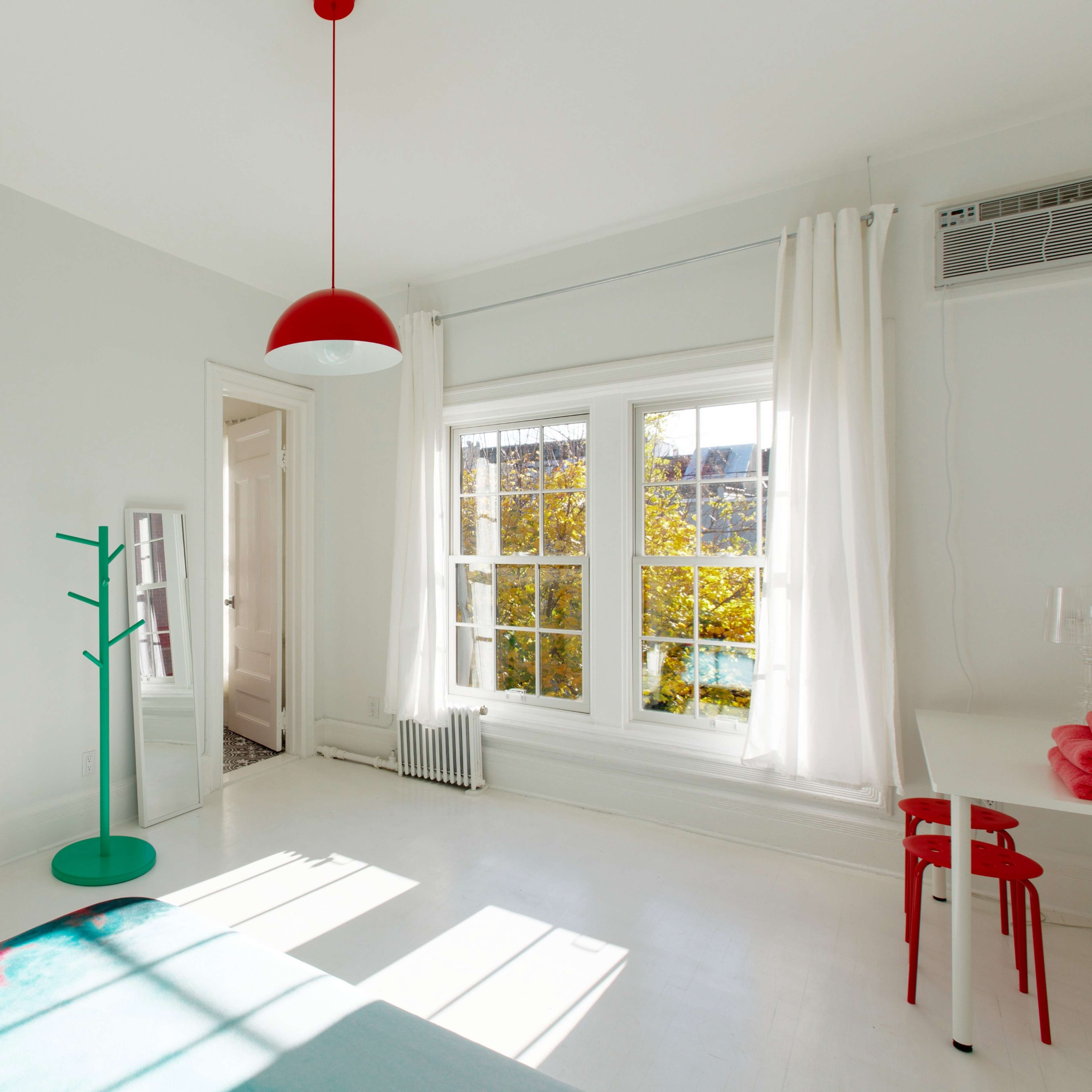 A bright, minimalistic bedroom with a white color scheme, featuring a red pendant light, a green coat rack, and large windows that let in natural light.