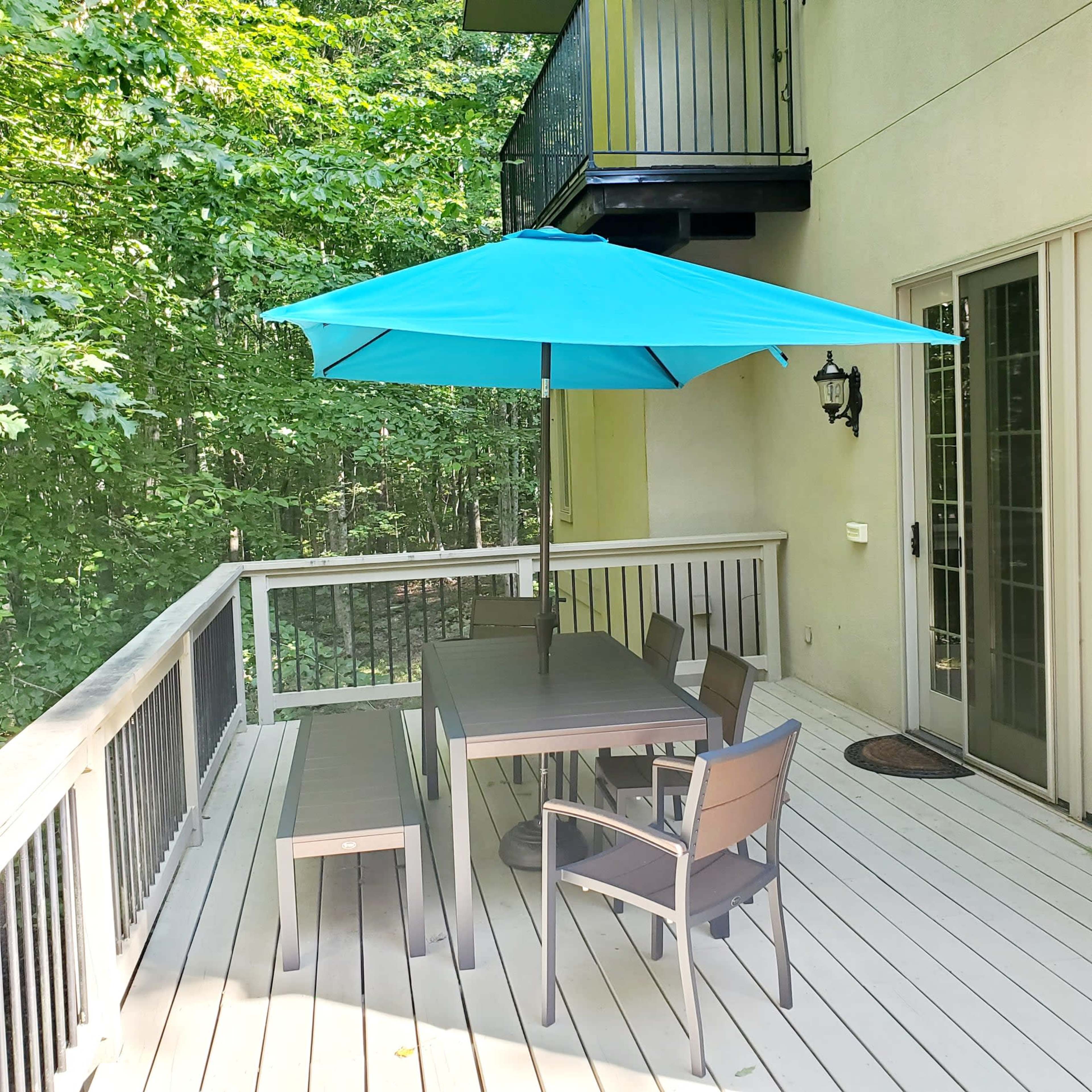 A deck area features a table with chairs and a bench, sheltered by a blue umbrella, surrounded by greenery.