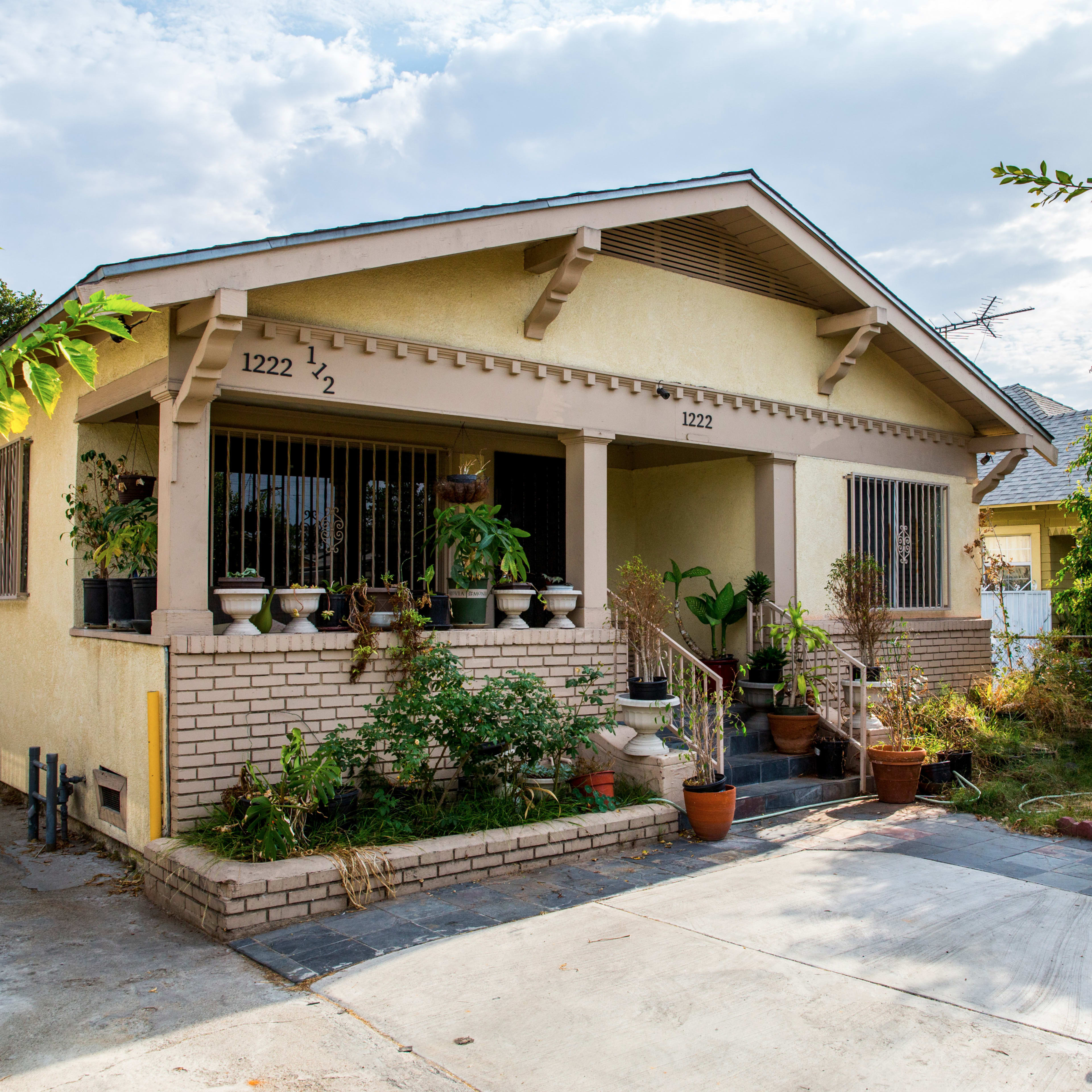 A single-story house with a covered porch, surrounded by various potted plants and trees, and a driveway leading to the front door.