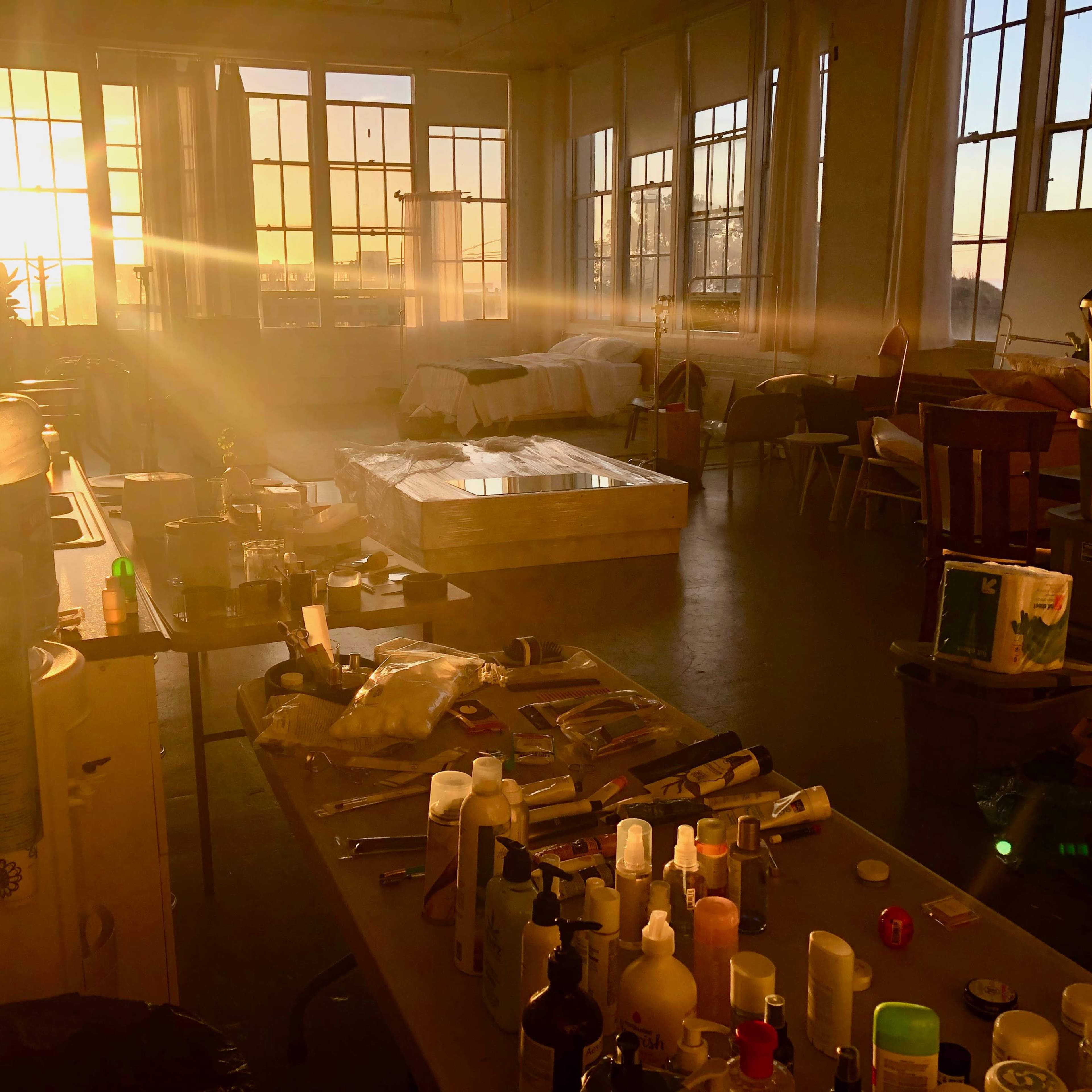 A sunlit room with large windows, containing a table cluttered with various bottles and materials, and a mattress in the background.