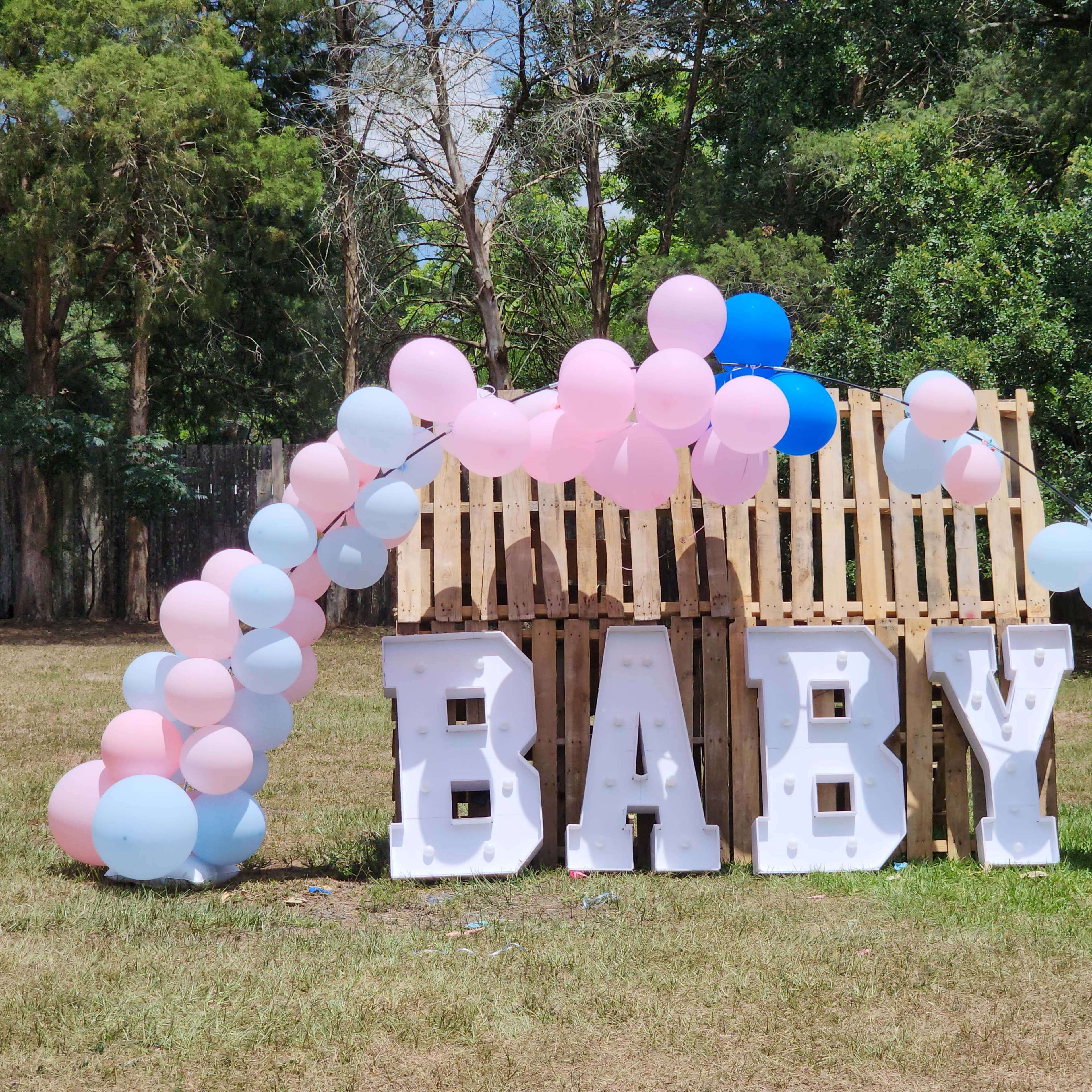 A decorative display featuring large white letters spelling "BABY" with an arch of pink and blue balloons in a grassy area.