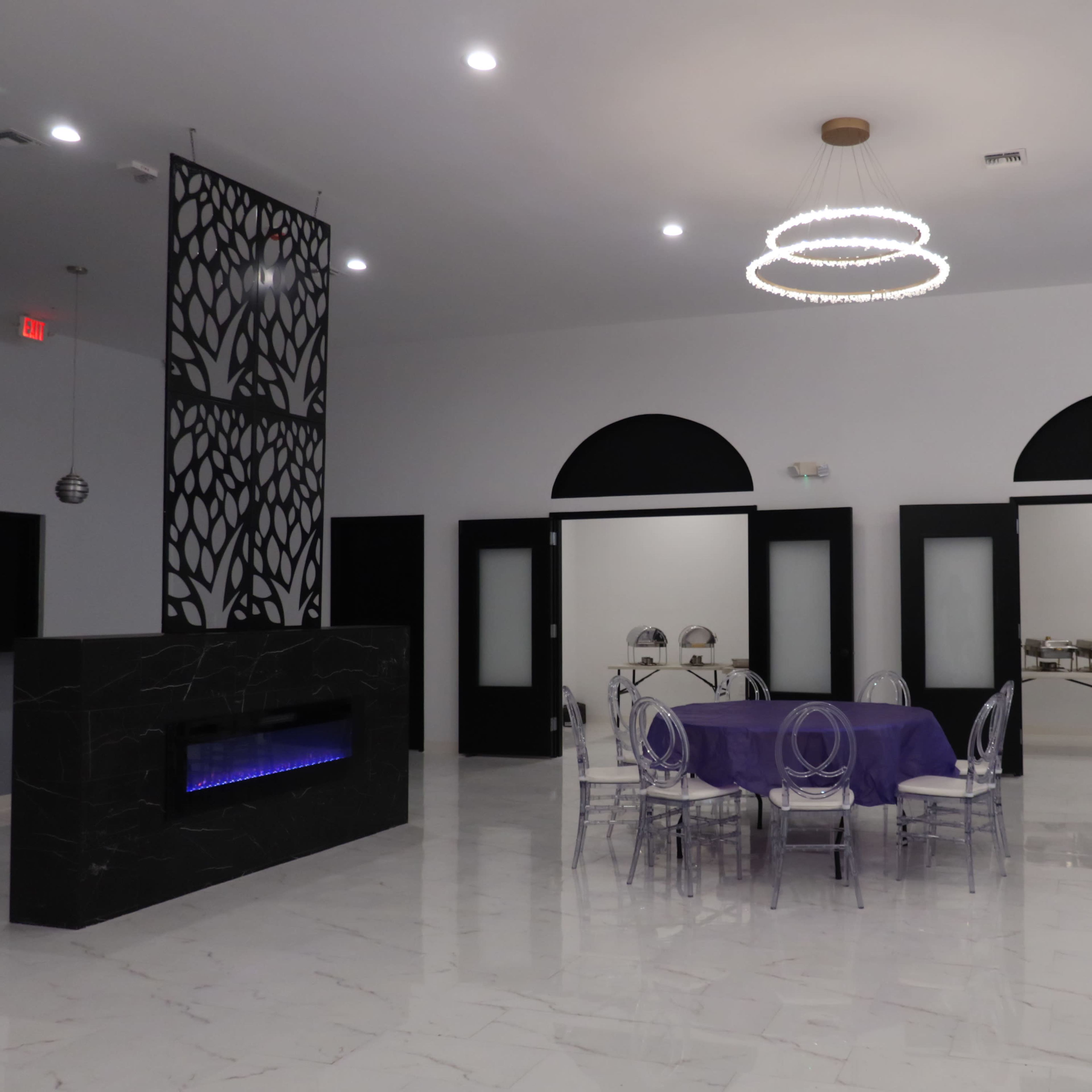 An elegant event space featuring purple tablecloths on round tables, clear acrylic chairs, a modern fireplace, and a decorative wall with a black design.