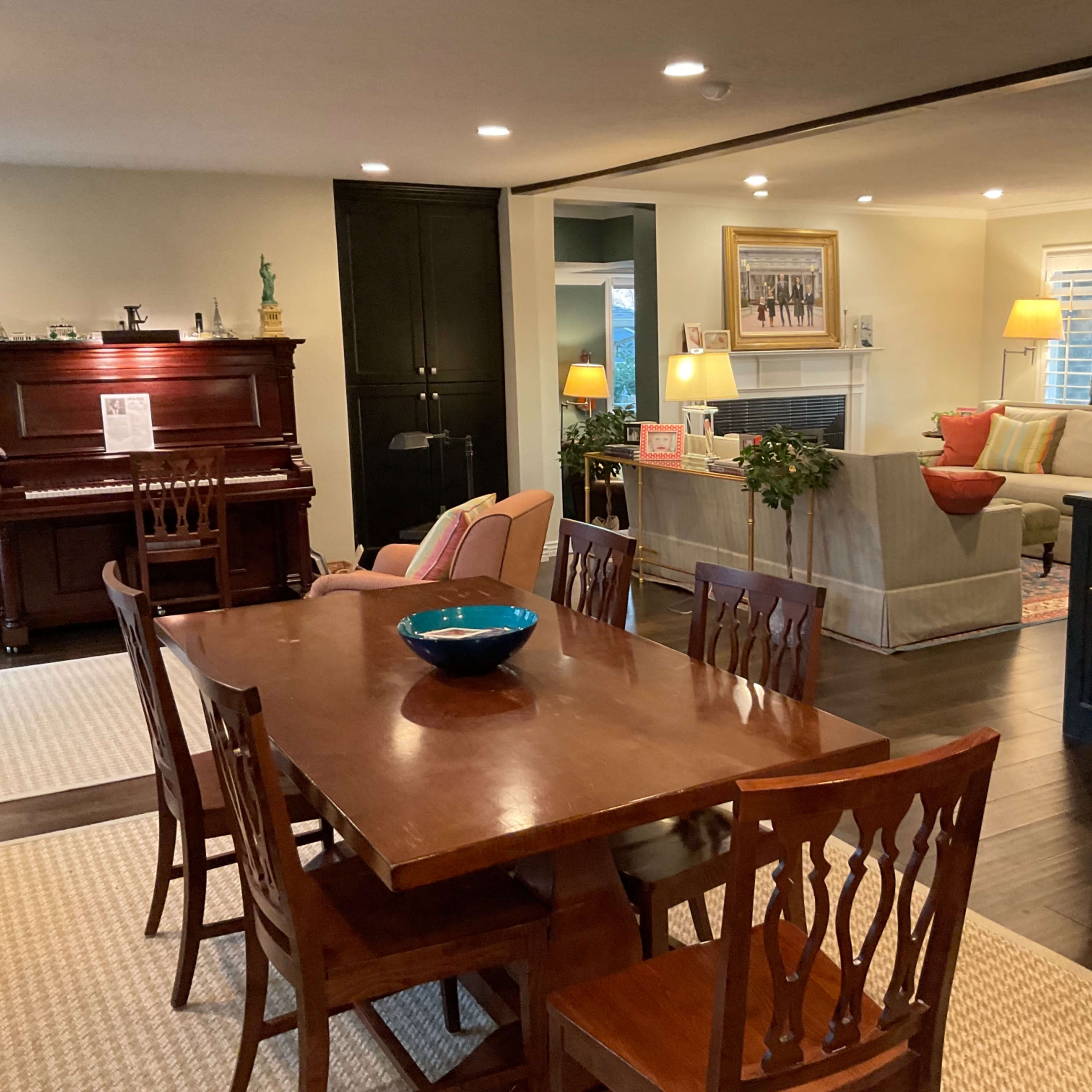 A dining area with a wooden table and chairs is situated near a living room that features a piano, plants, and soft lighting.