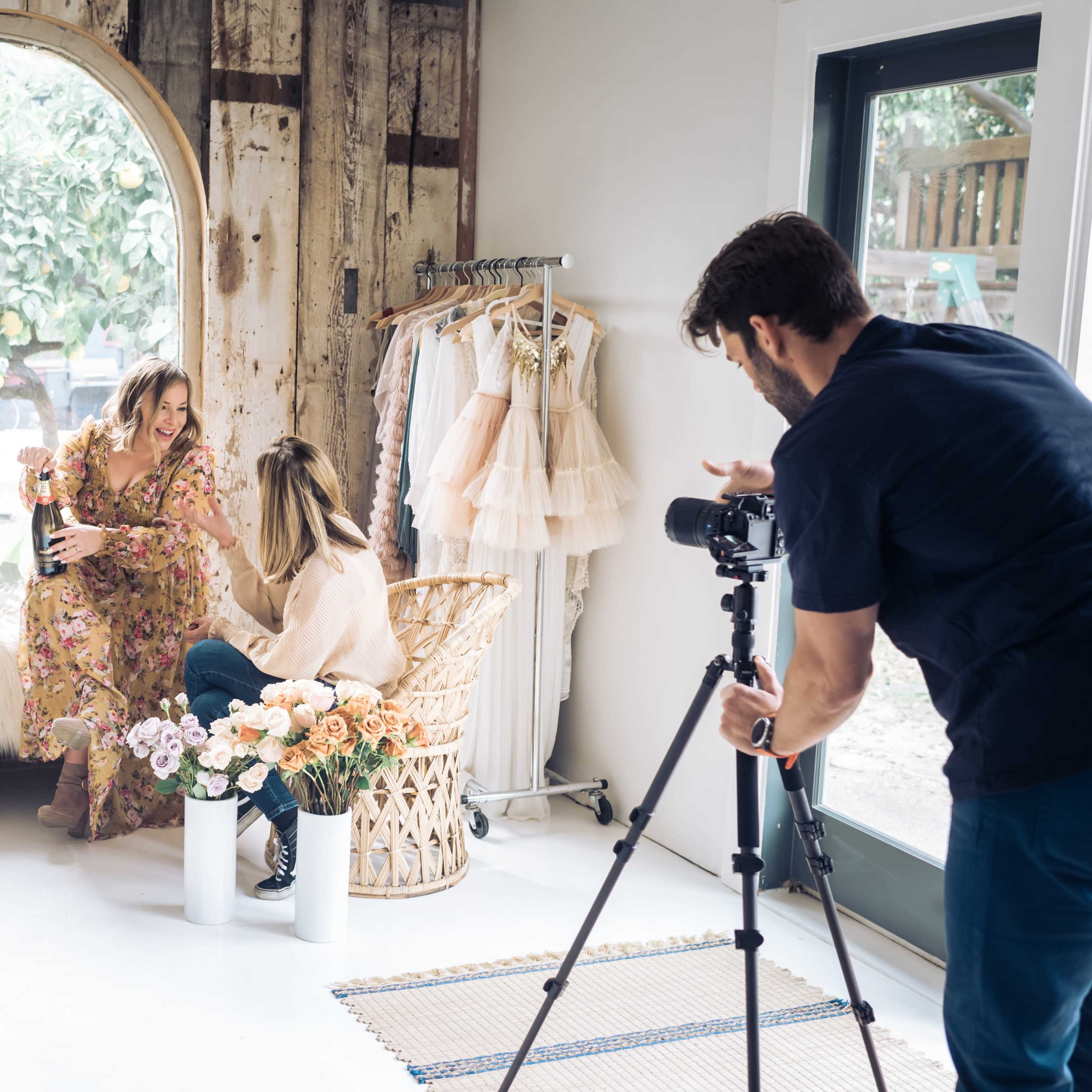 A group of four women sits and interacts while a man films them with a camera in a cozy, stylish studio setting.