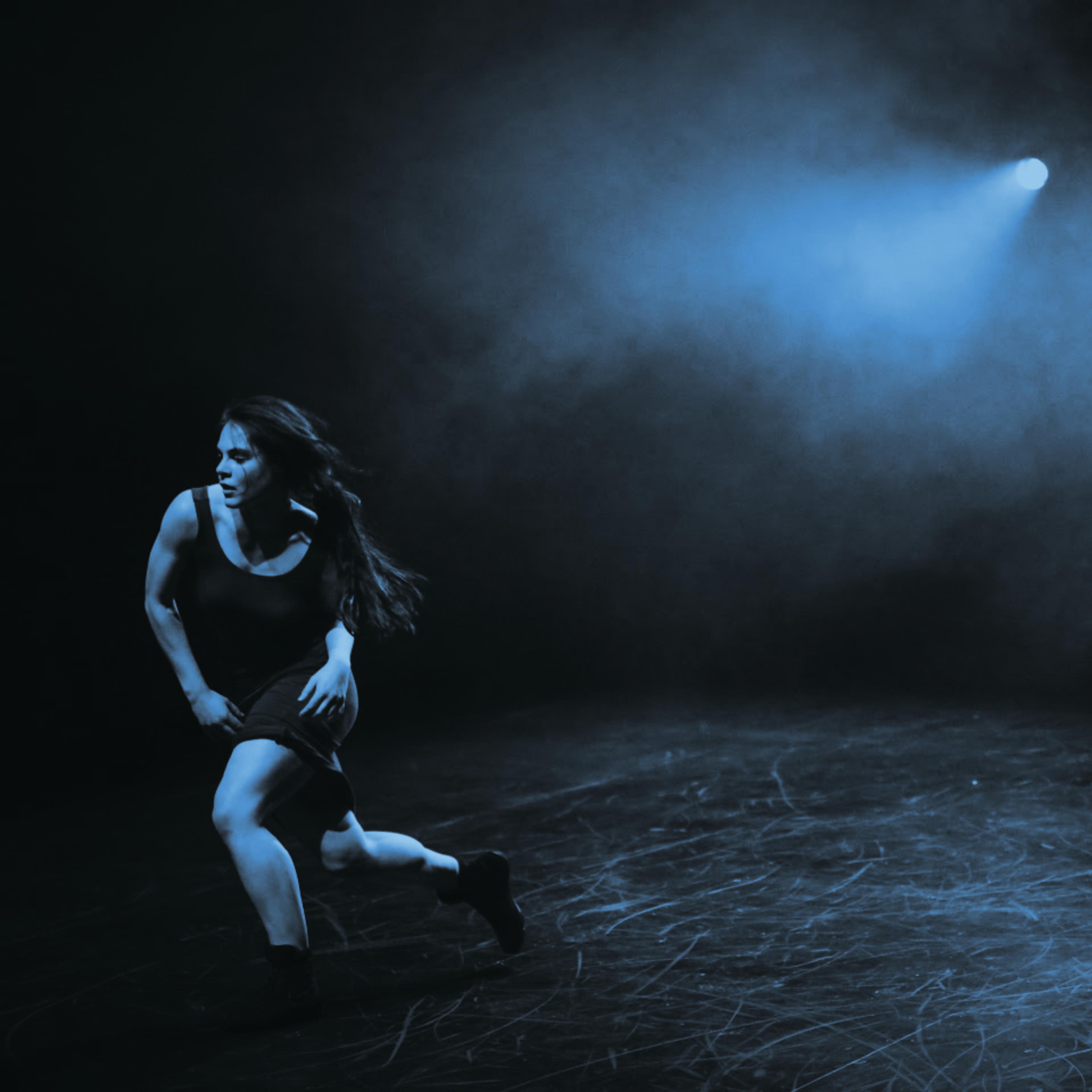 A dancer moves across a dimly lit stage, illuminated by blue spotlight beams amidst a smoky backdrop.