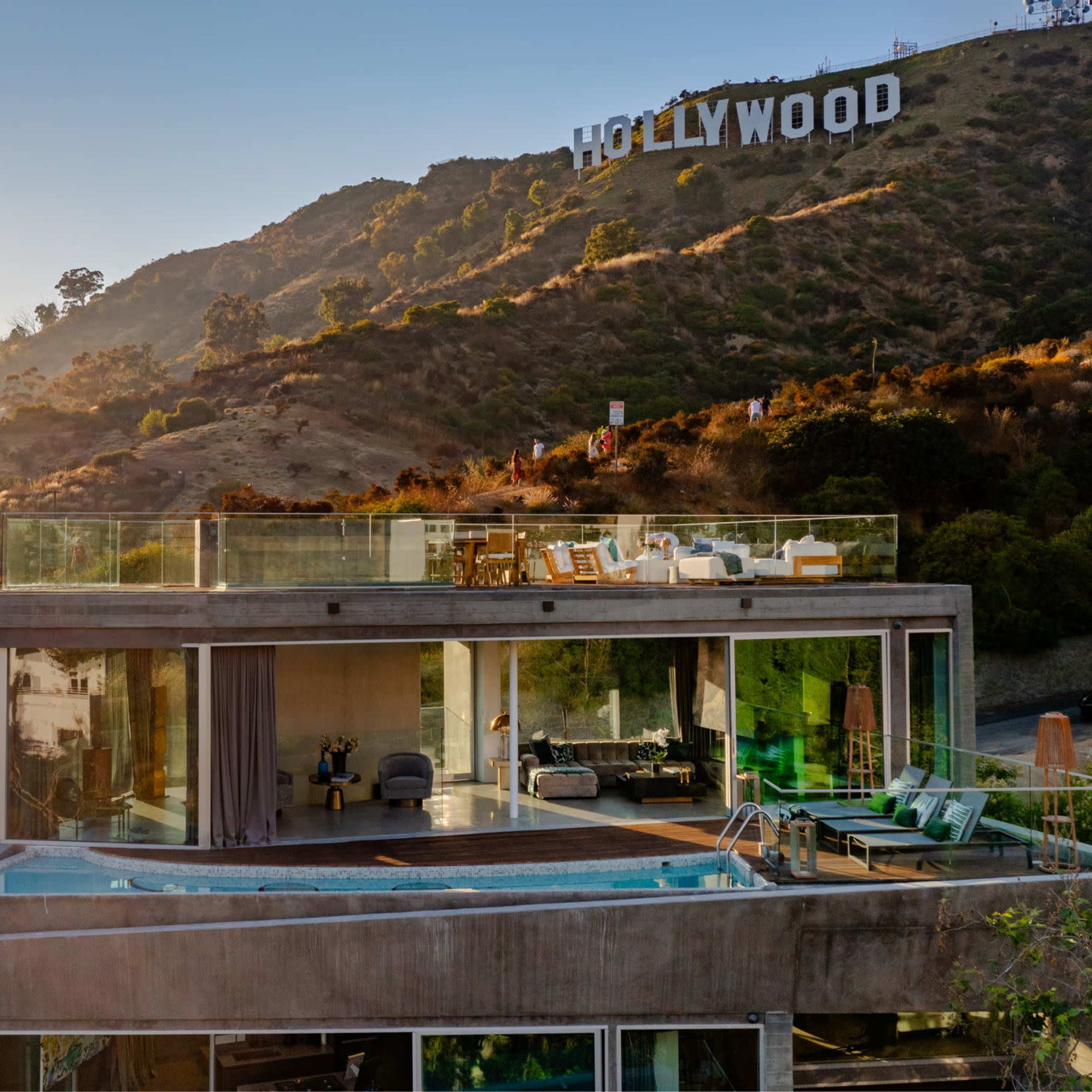 A modern, multi-level house with a glass facade features a rooftop pool and overlooks the Hollywood sign on a hillside.