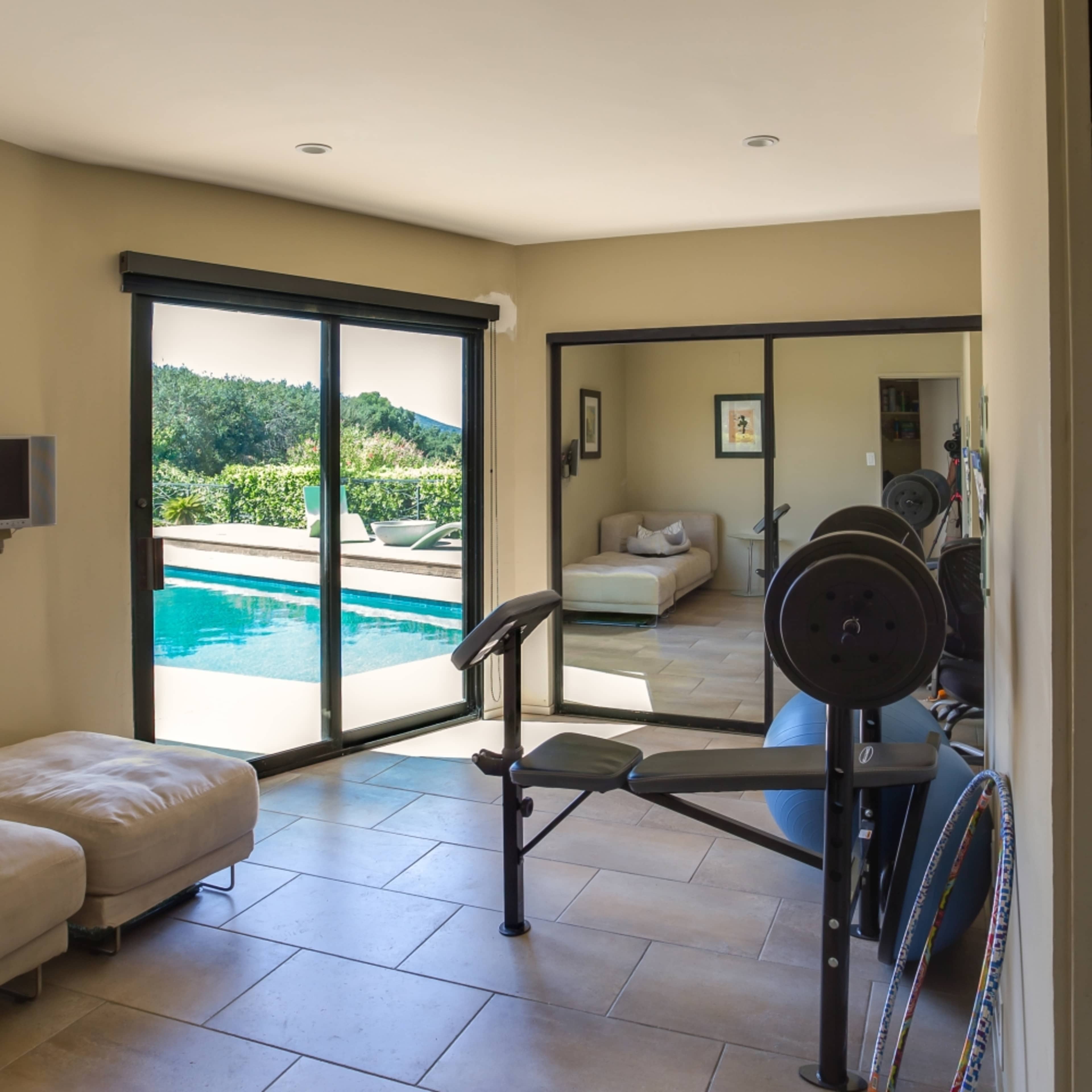 A home gym area features a weight bench and exercise equipment, with large windows overlooking a swimming pool and outdoor seating.