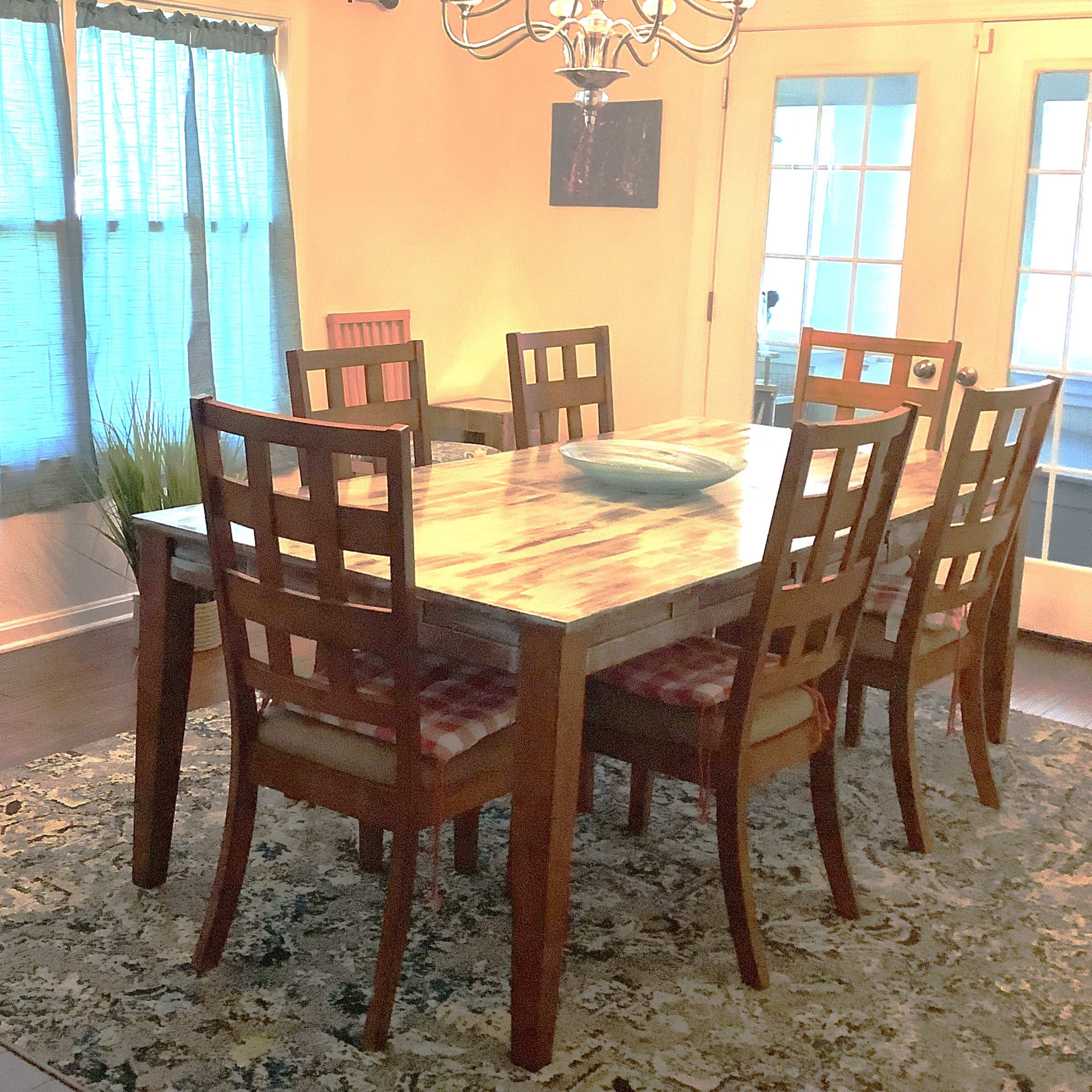 A wooden dining table with eight chairs is set in a well-lit room featuring large glass doors and blue curtains.