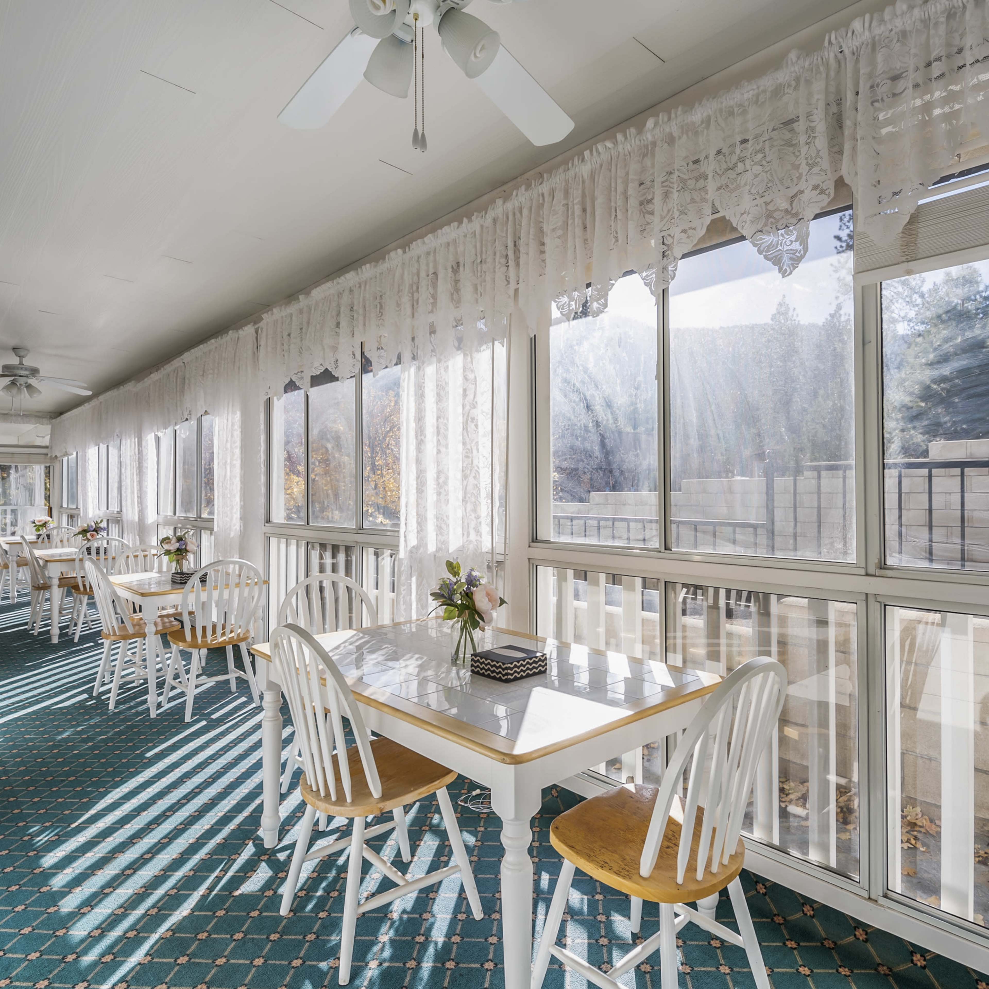 A sunlit sunroom features white tables and chairs along its length, with lace curtains and a patterned carpet.