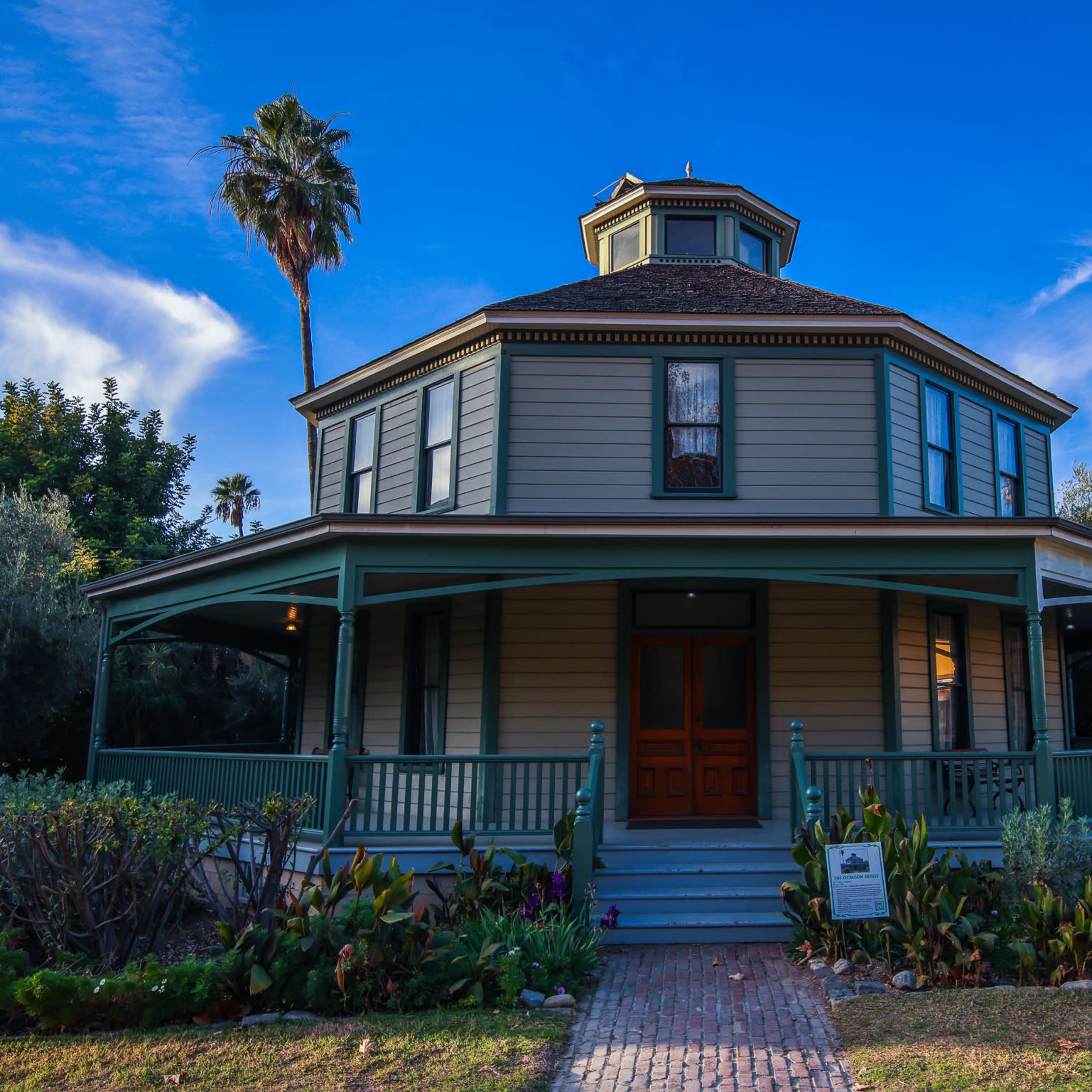 A large, round, two-story house with a green porch, surrounded by plants and trees under a blue sky.