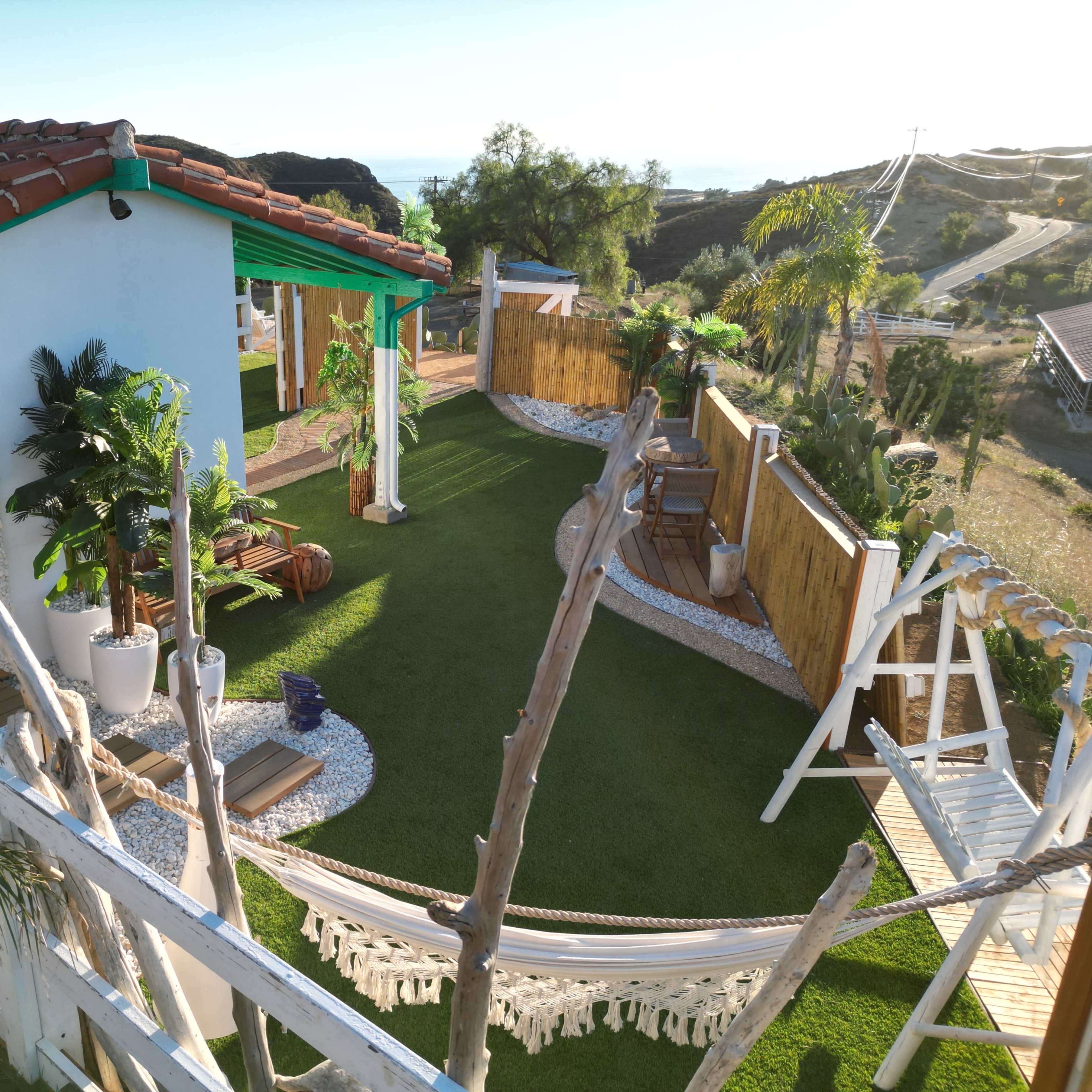 A landscape view of a landscaped backyard featuring a hammock, seating areas, greenery, and a pathway leading to a hillside.