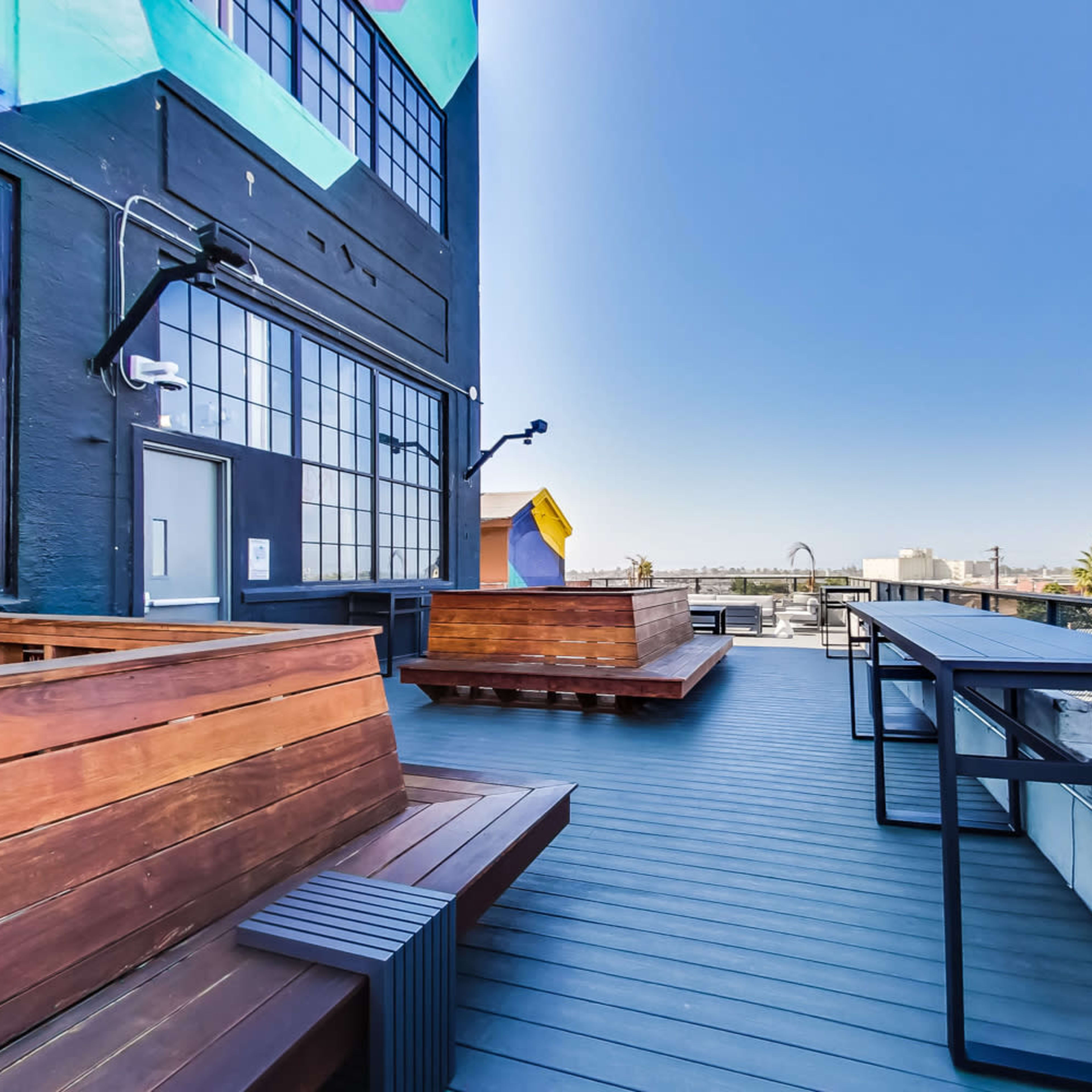 A rooftop area with wooden seating and tables, overlooking a clear sky and distant structures.