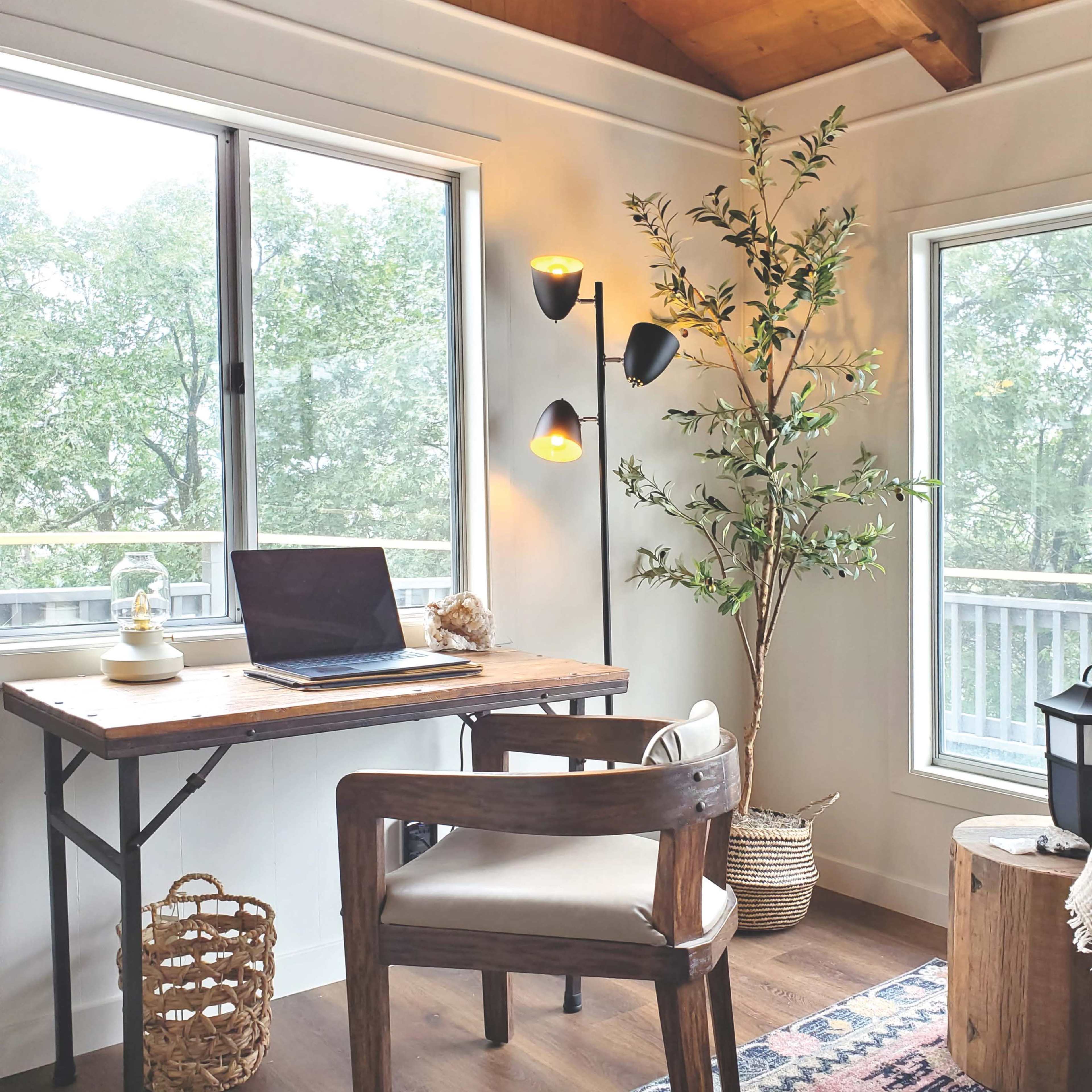 A workspace with a wooden desk, a laptop, a chair, and a potted plant, located near large windows with natural light.