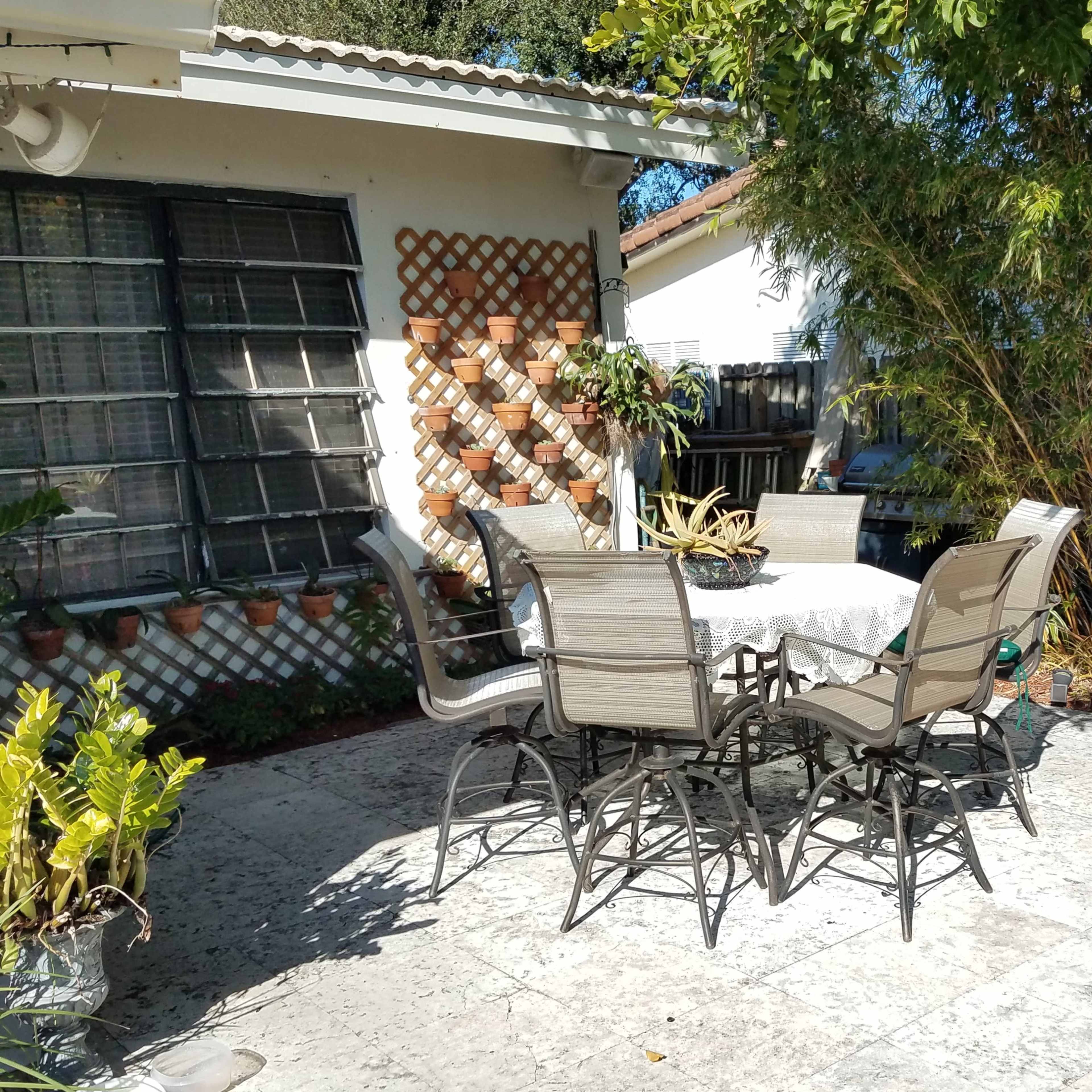 A stone patio with a table and six chairs surrounded by plants and a house in the background.