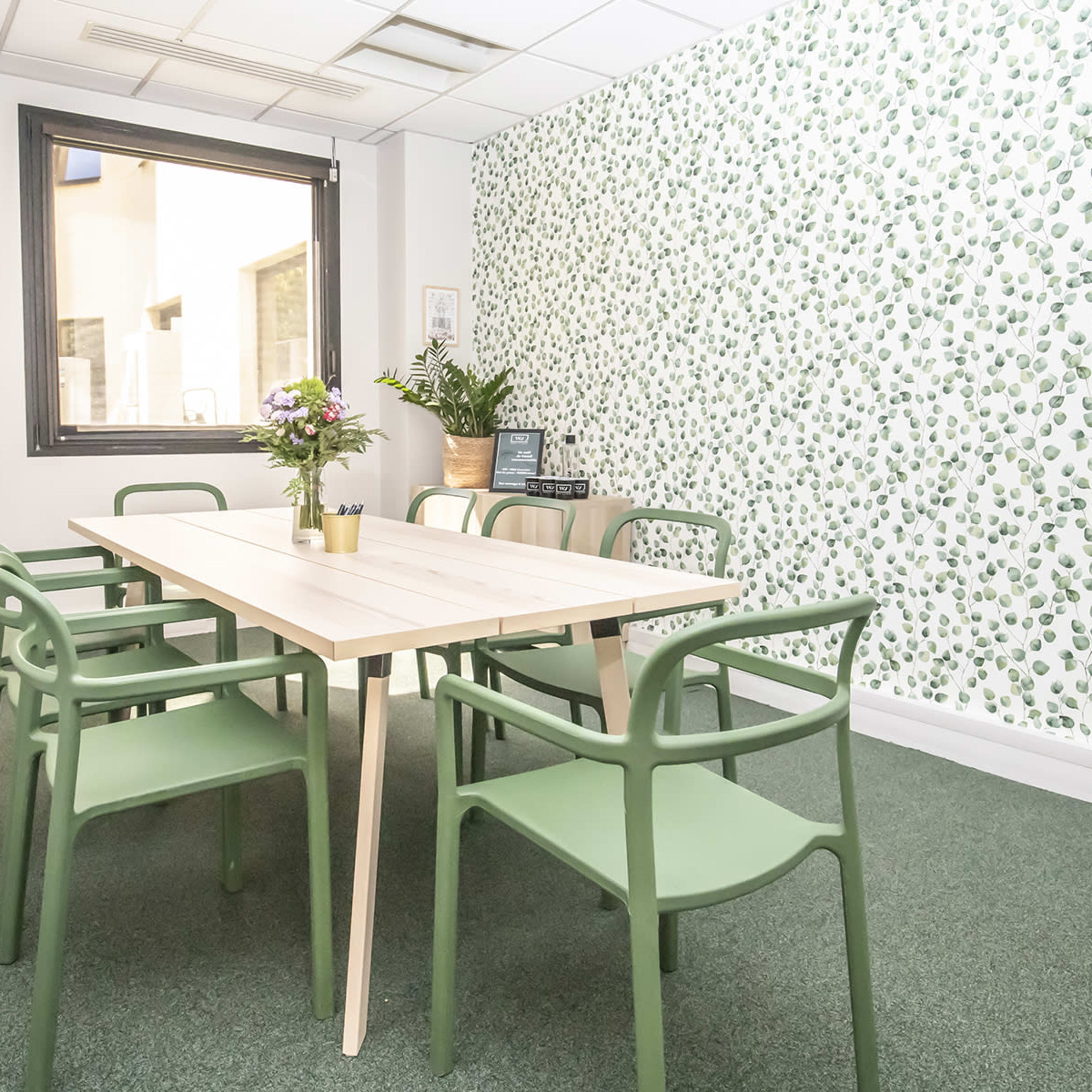 A bright meeting room with a large wooden table surrounded by green chairs, and a wall decorated with a leafy pattern.