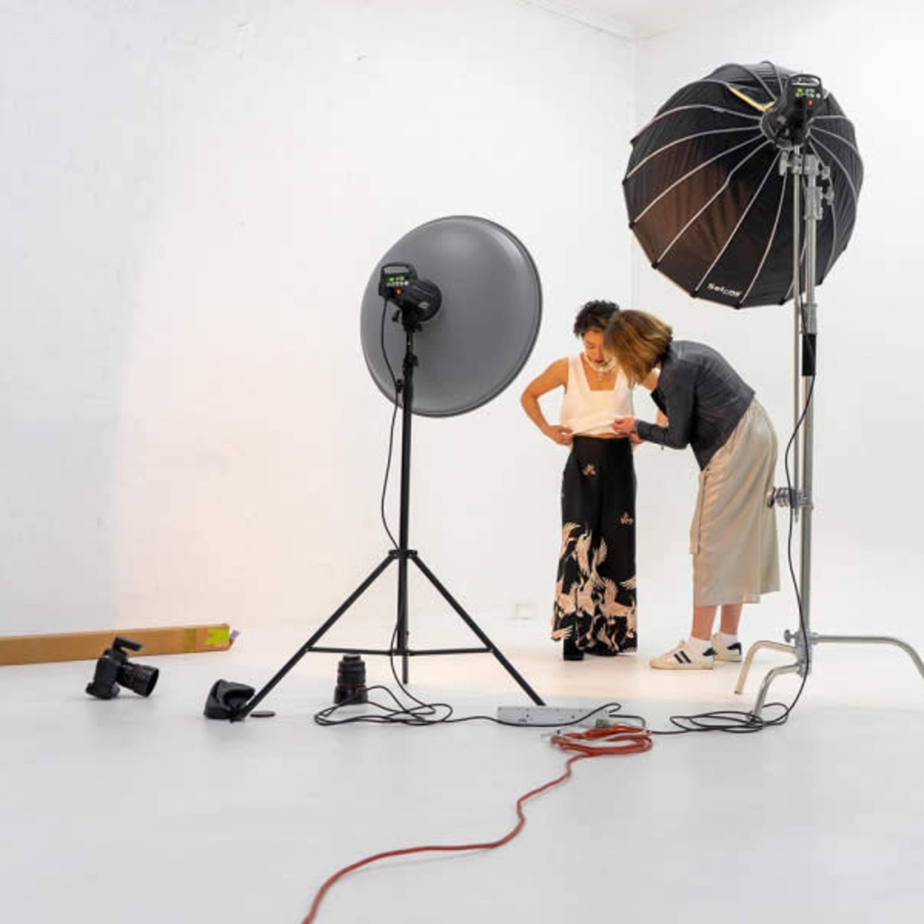 Two women are adjusting clothing in a well-lit photography studio featuring large softbox lights and a seamless white backdrop.