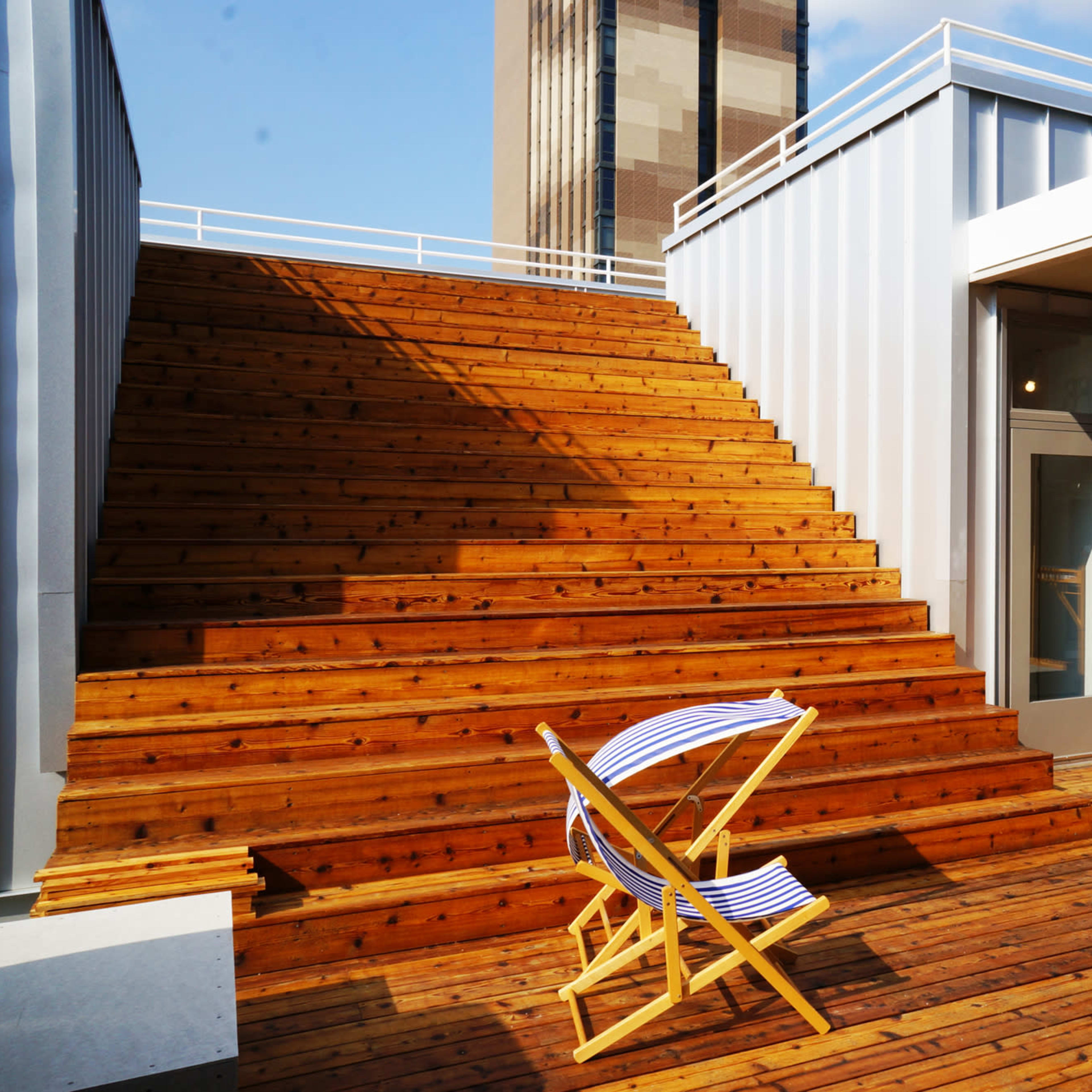 A wooden staircase leading up to a rooftop area, flanked by modern metal structures and featuring a single deck chair on the wooden floor.