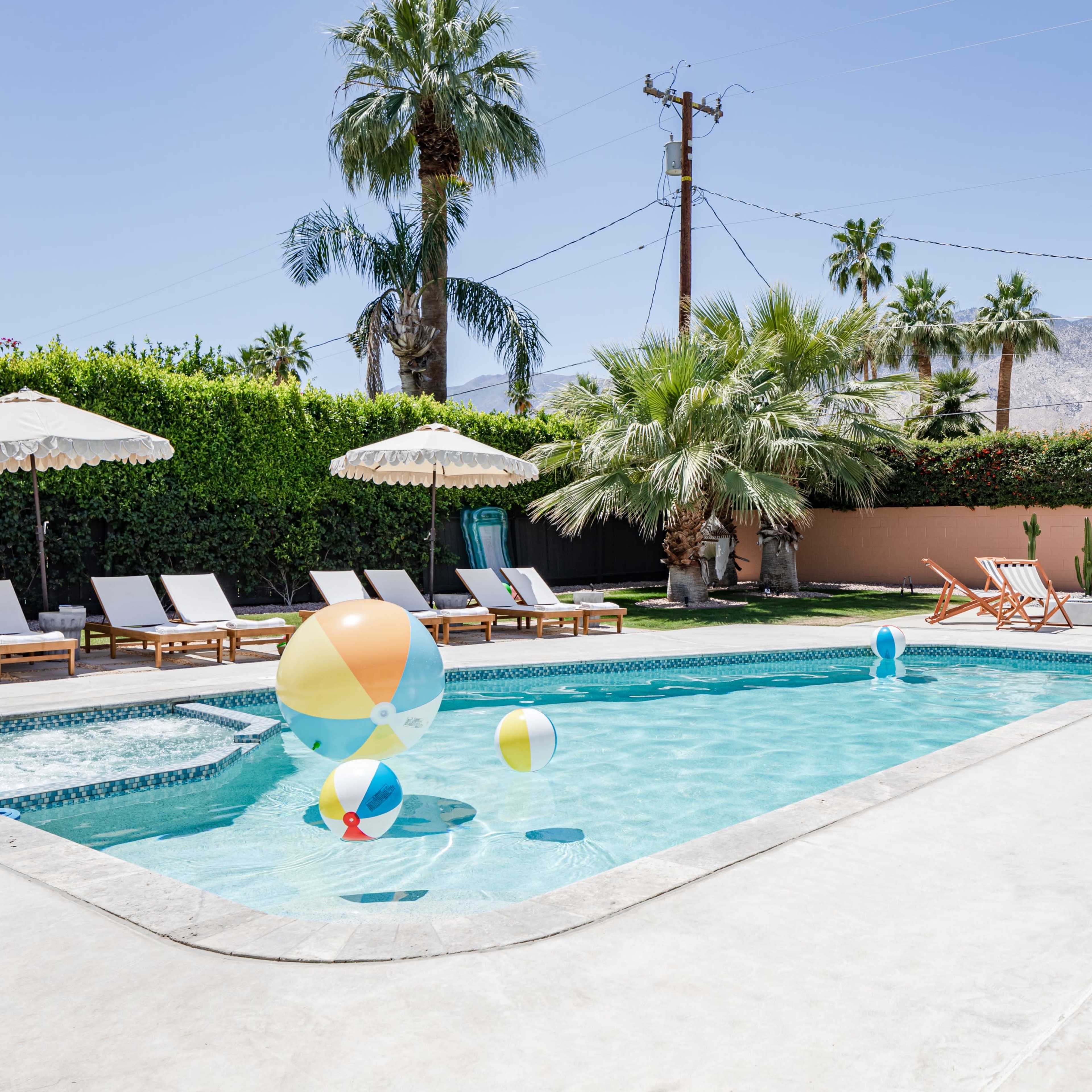 A sparkling swimming pool surrounded by lounge chairs and beach balls, with tall palm trees in the background.