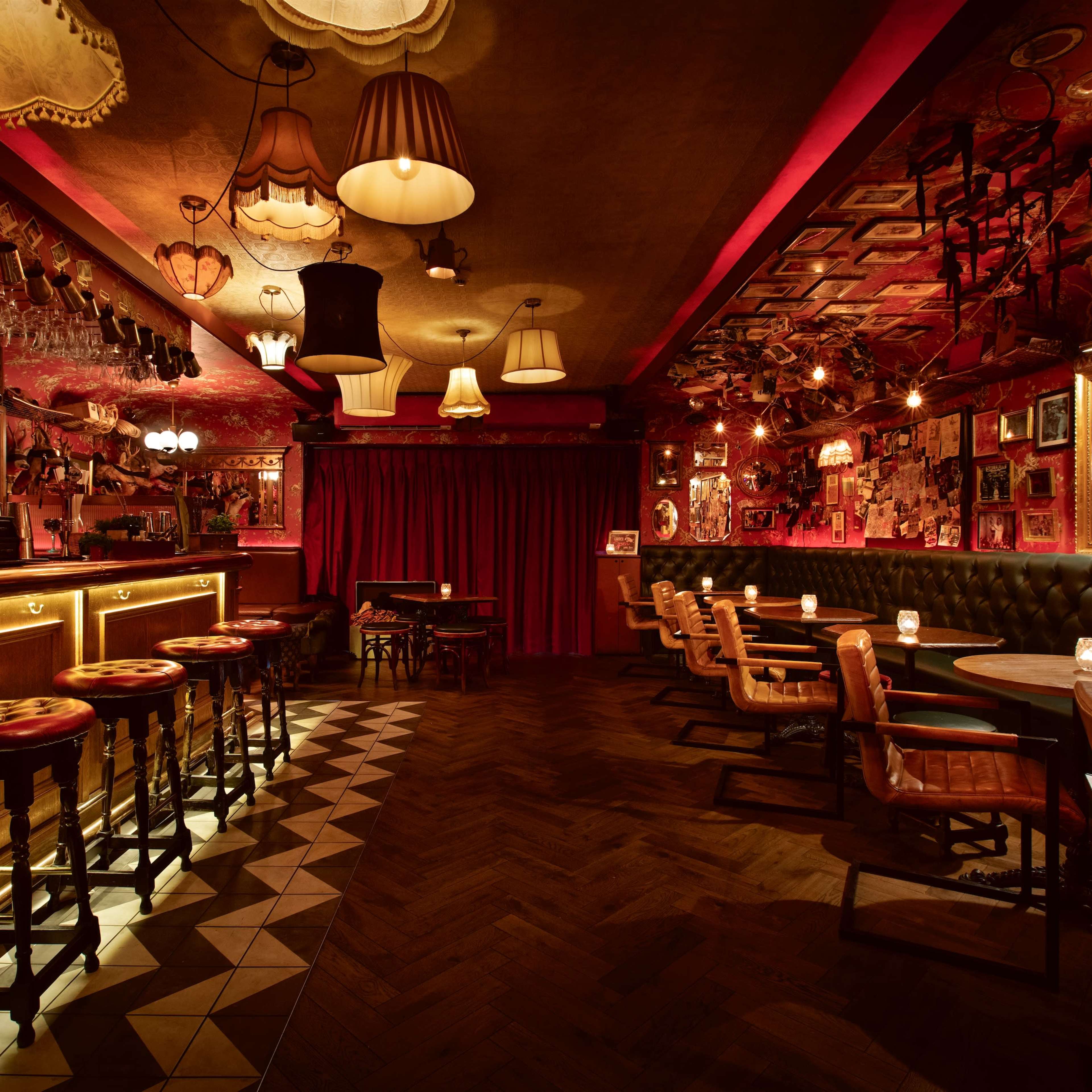 A dimly lit bar with wooden stools, a long counter lined with bottles, and diverse seating arrangements beneath decorative lamps and framed artwork.