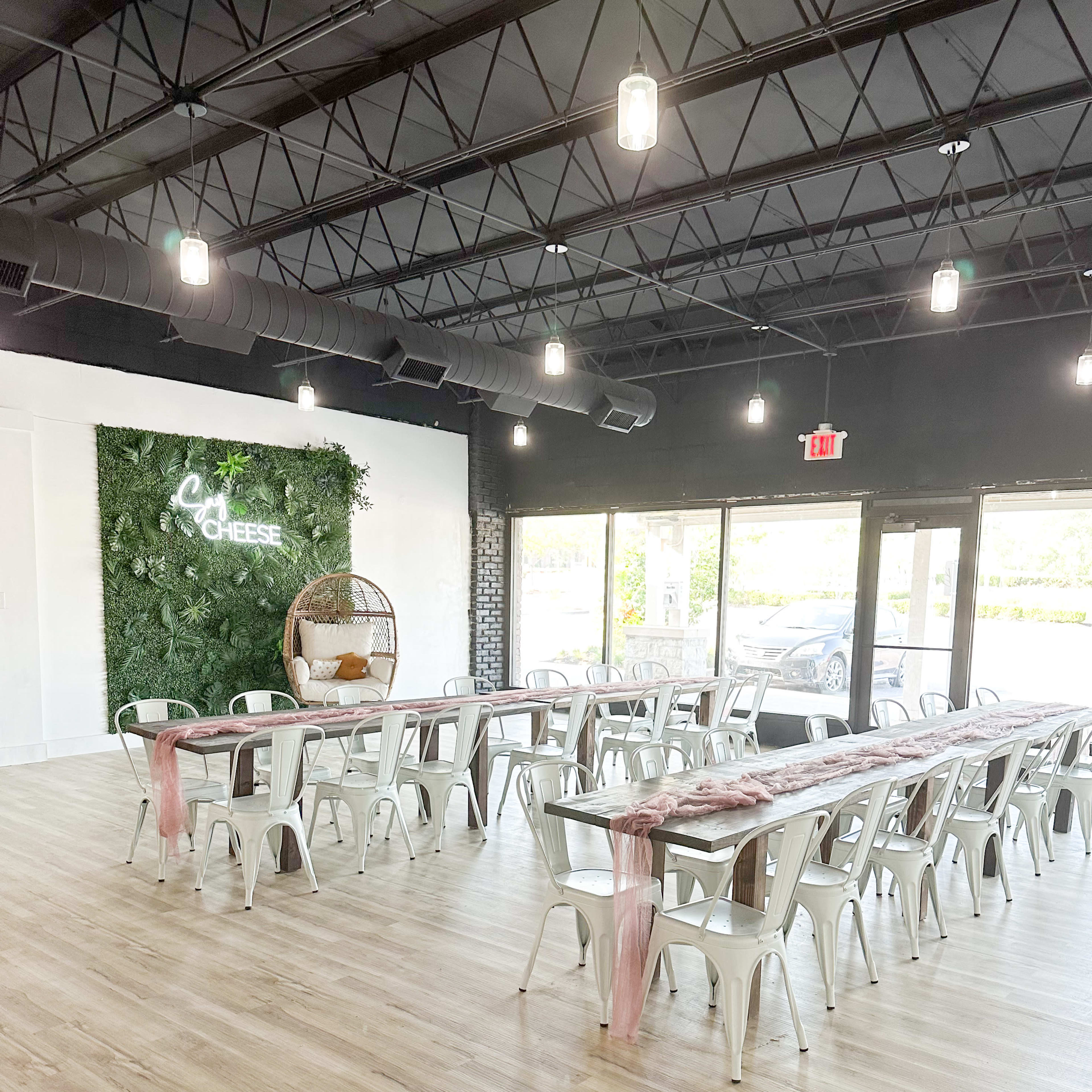 A spacious event venue with long tables arranged in rows, decorated with pink runners, and a green wall featuring the words "Cheese."