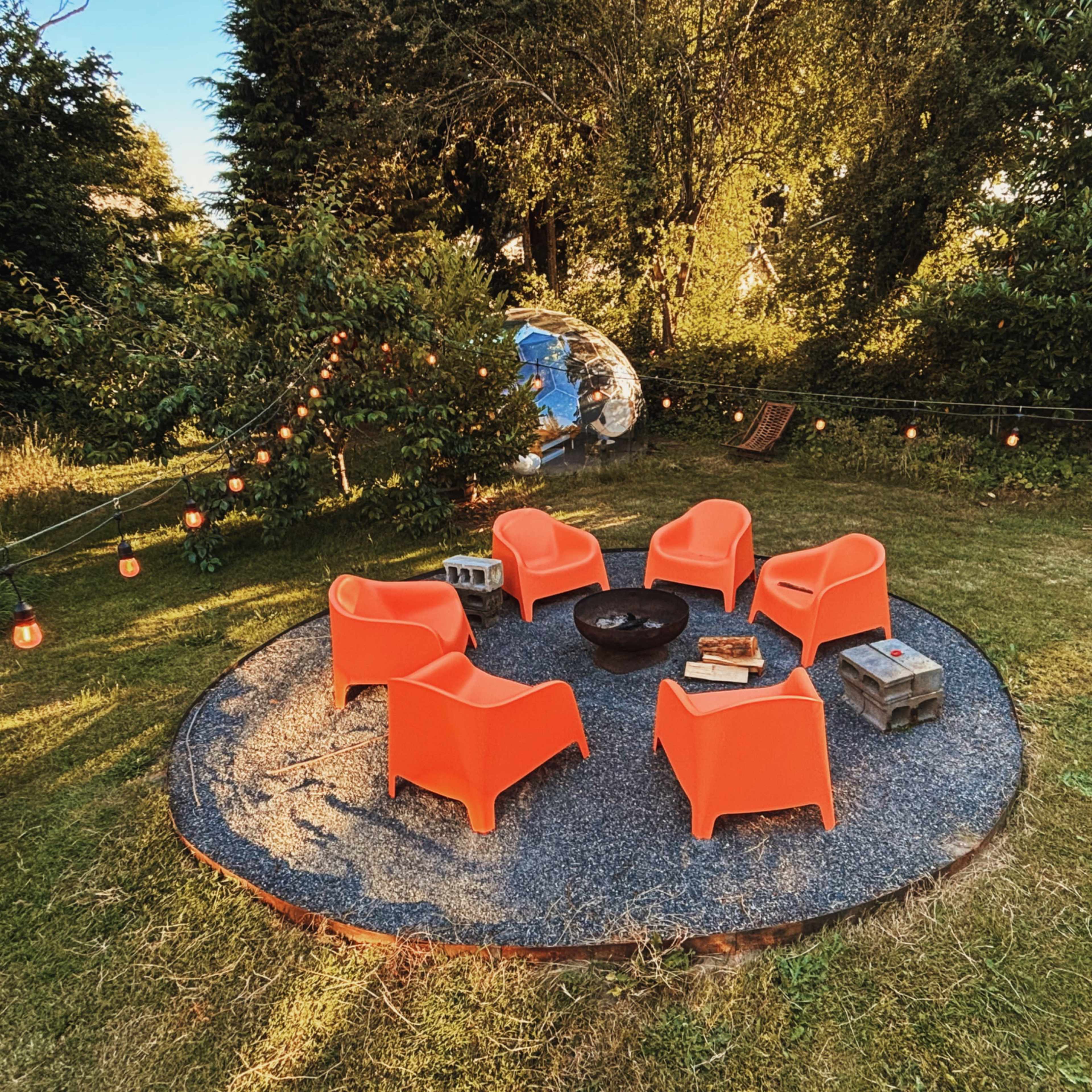 A circular arrangement of five orange chairs surrounds a fire pit on a gravel patio in a grassy yard illuminated by string lights.