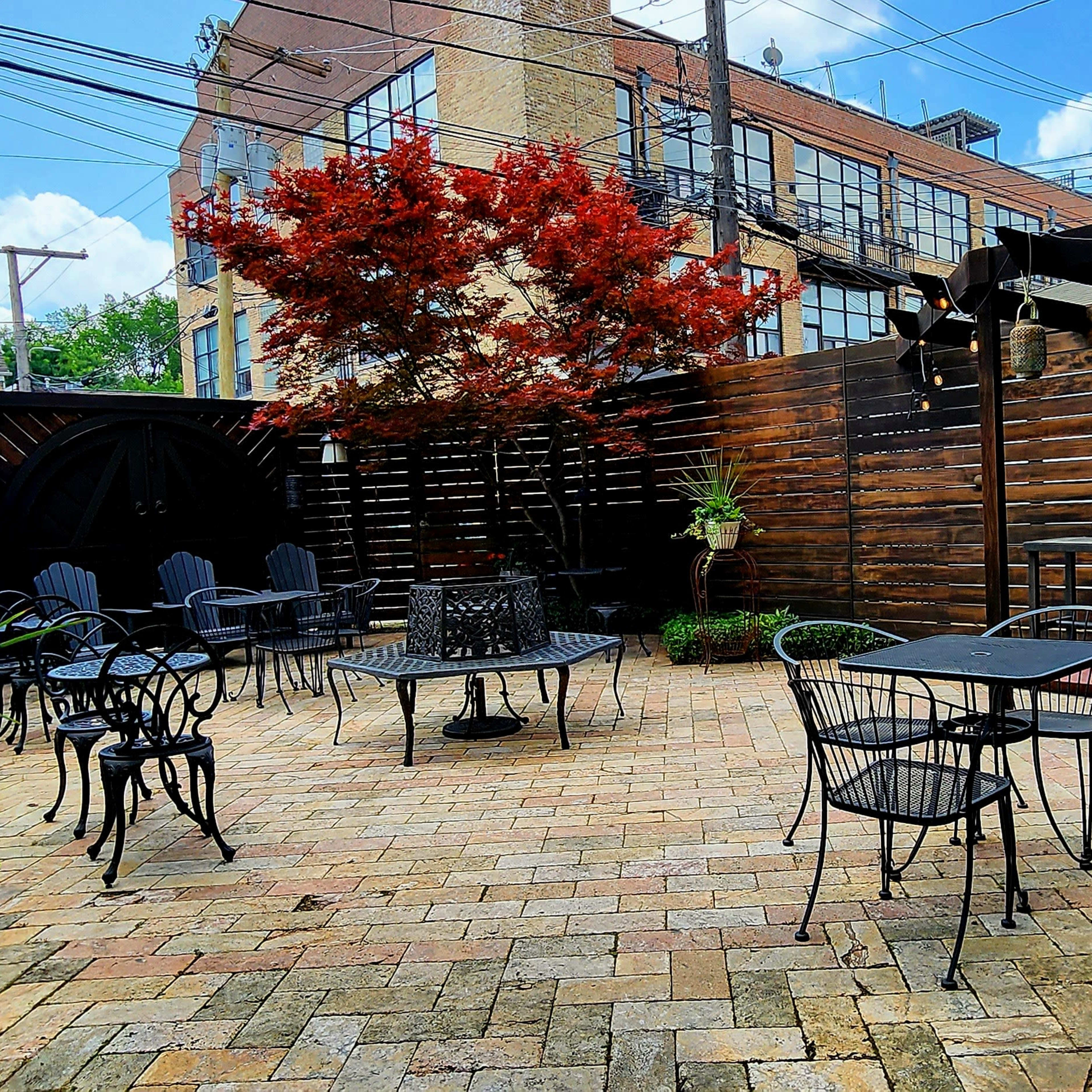 A brick-paved patio features multiple metal tables and chairs surrounded by a wooden fence and potted plants.