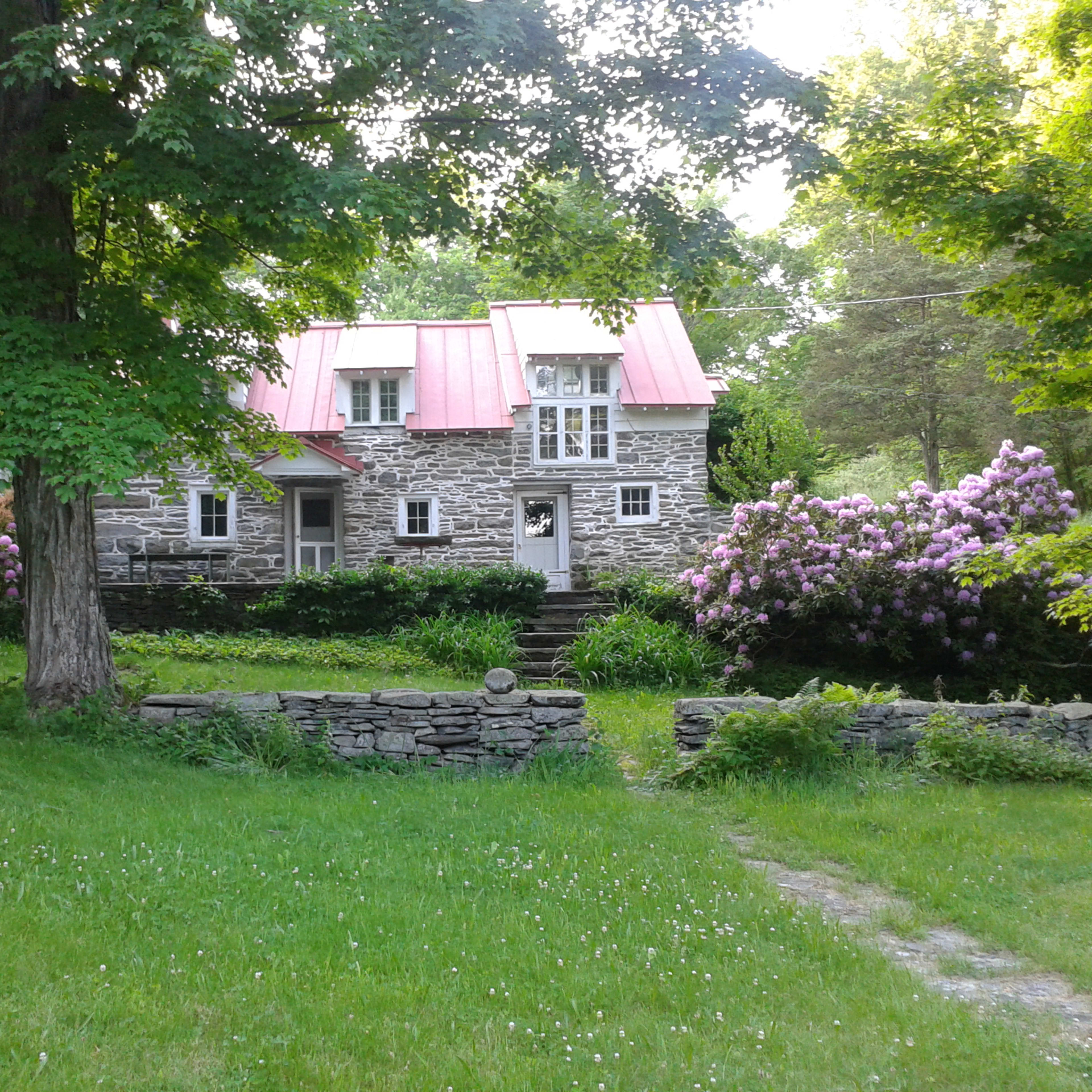 A stone house with a red metal roof is surrounded by green grass and blooming shrubs.