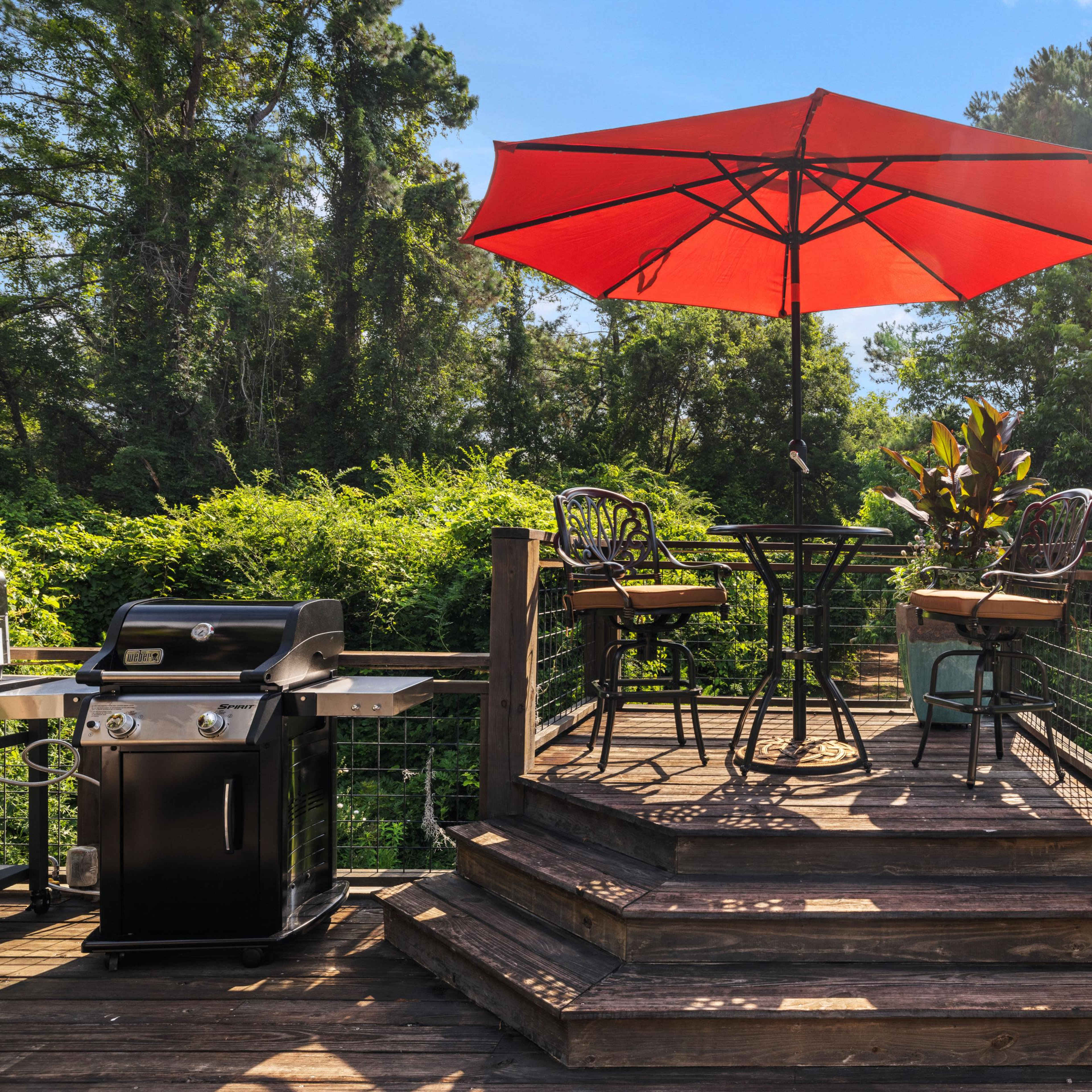 A circular wooden deck features a black metal table and chairs under a large red umbrella, with a grill nearby and a birdhouse on the railing.
