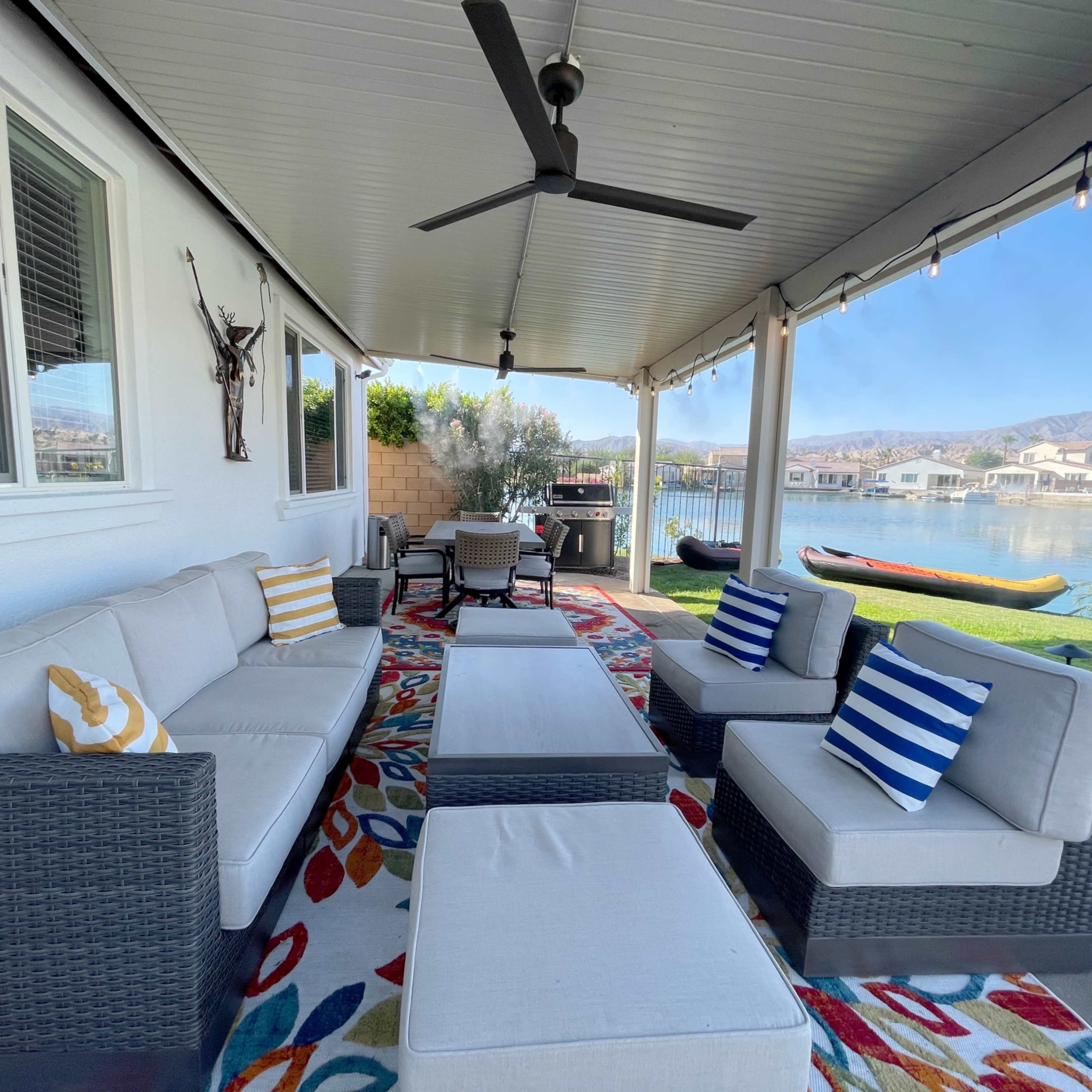 A covered patio area featuring a seating arrangement with a coffee table, decorative pillows, and a view of a lake and houses in the background.