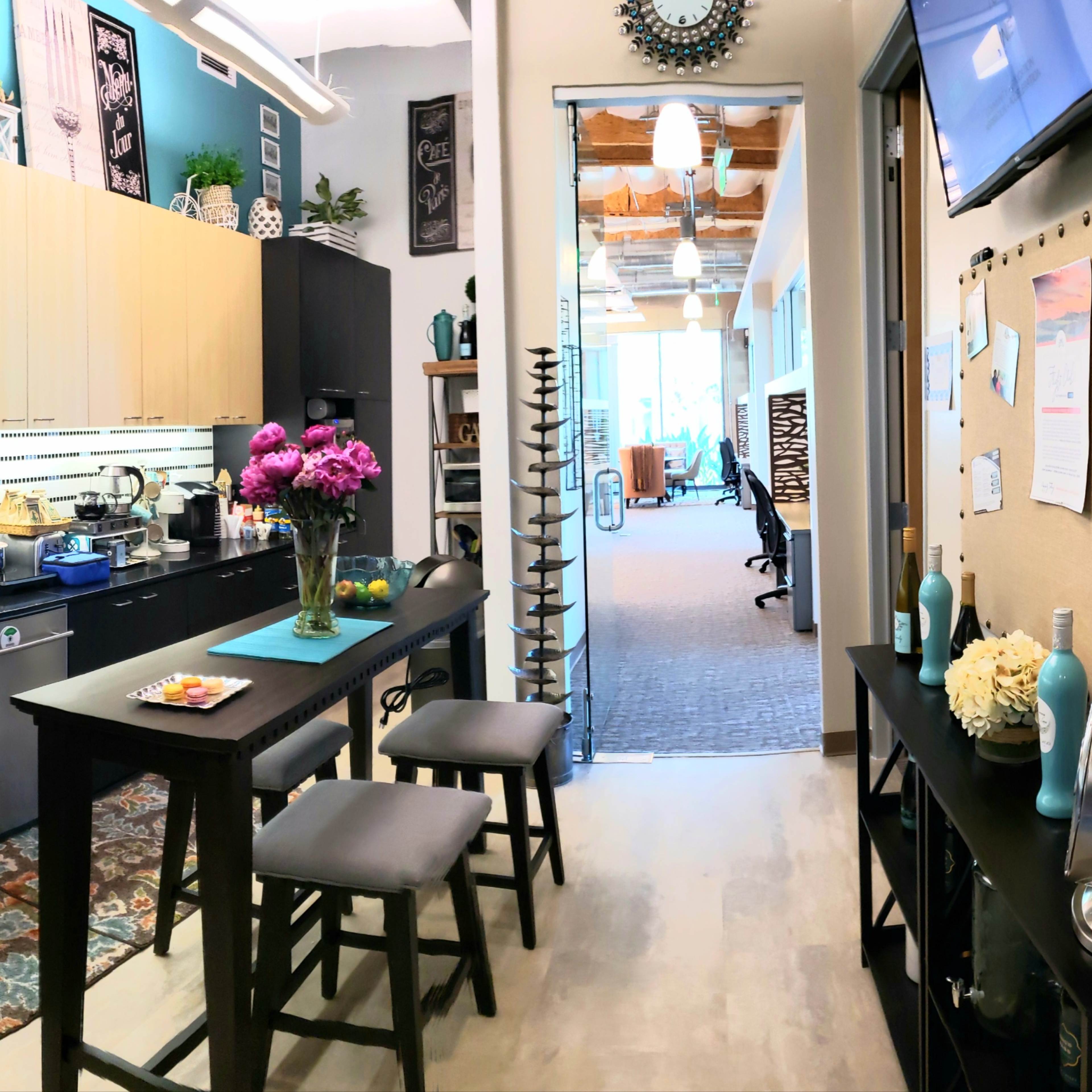 A modern kitchen area with a long table and stools, leading into an open workspace decorated with plants and artwork.
