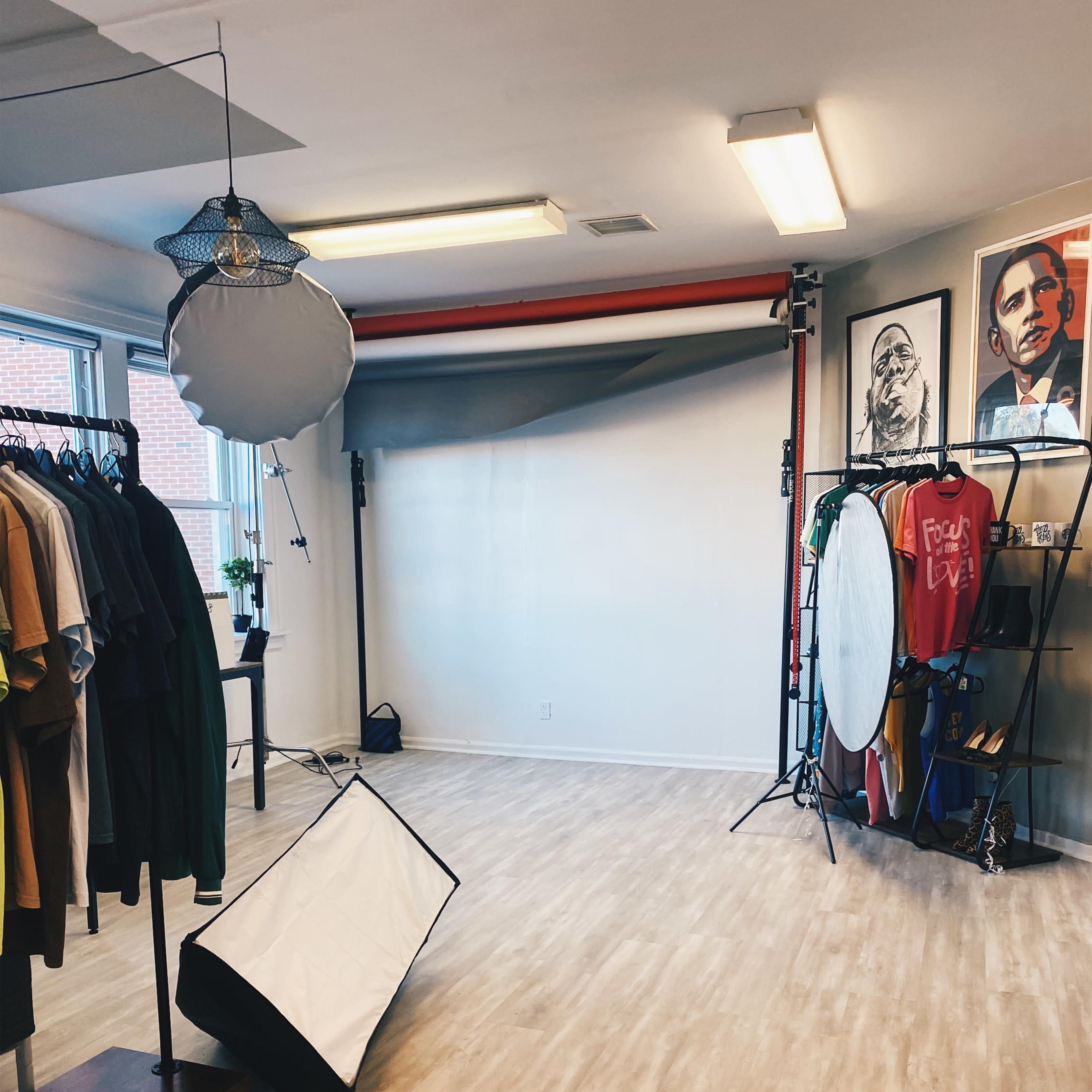 A photography studio featuring a backdrop, various lighting equipment, and clothing racks against light-colored walls.