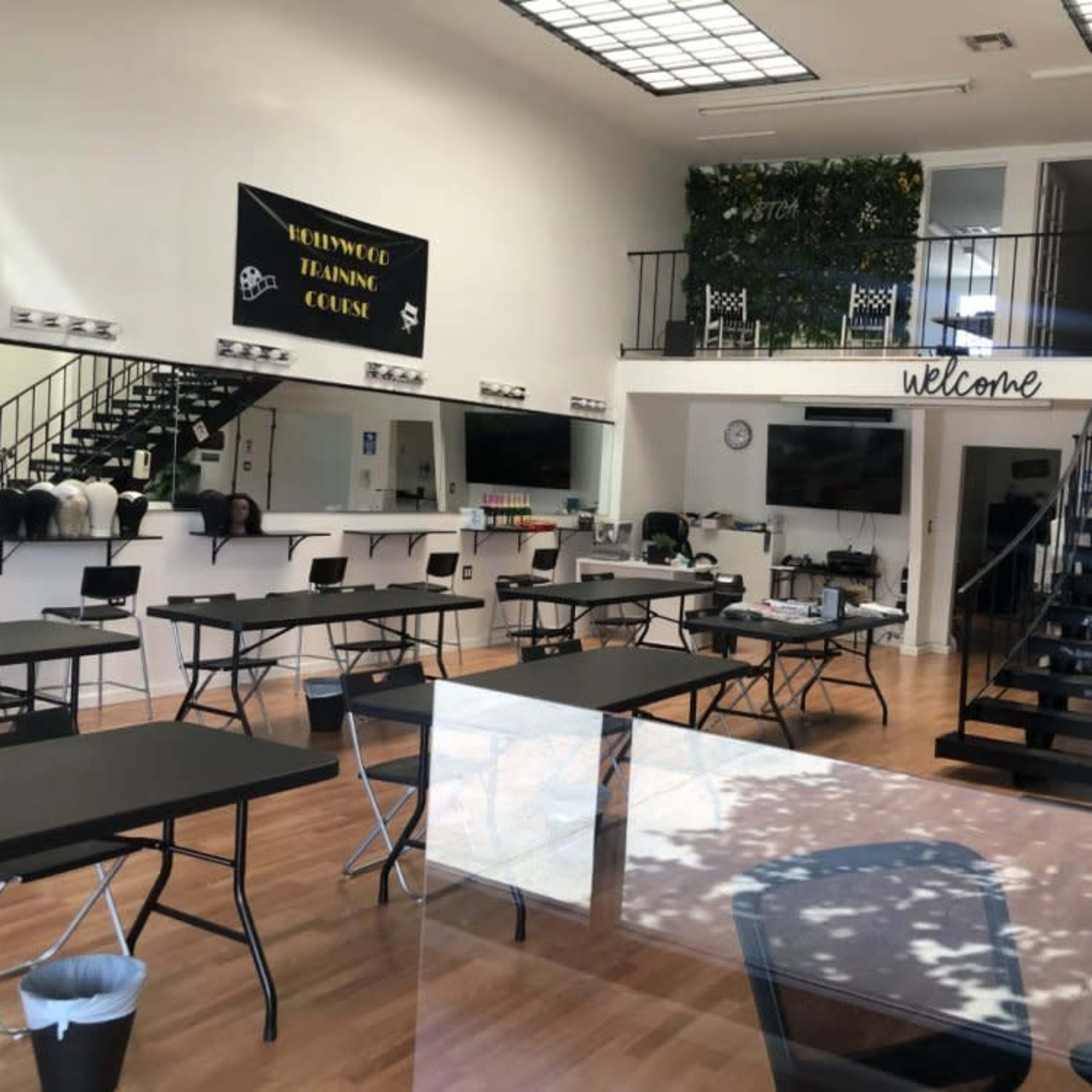 A spacious classroom setting with multiple tables arranged for a workshop, large windows, and a staircase leading to an upper level.
