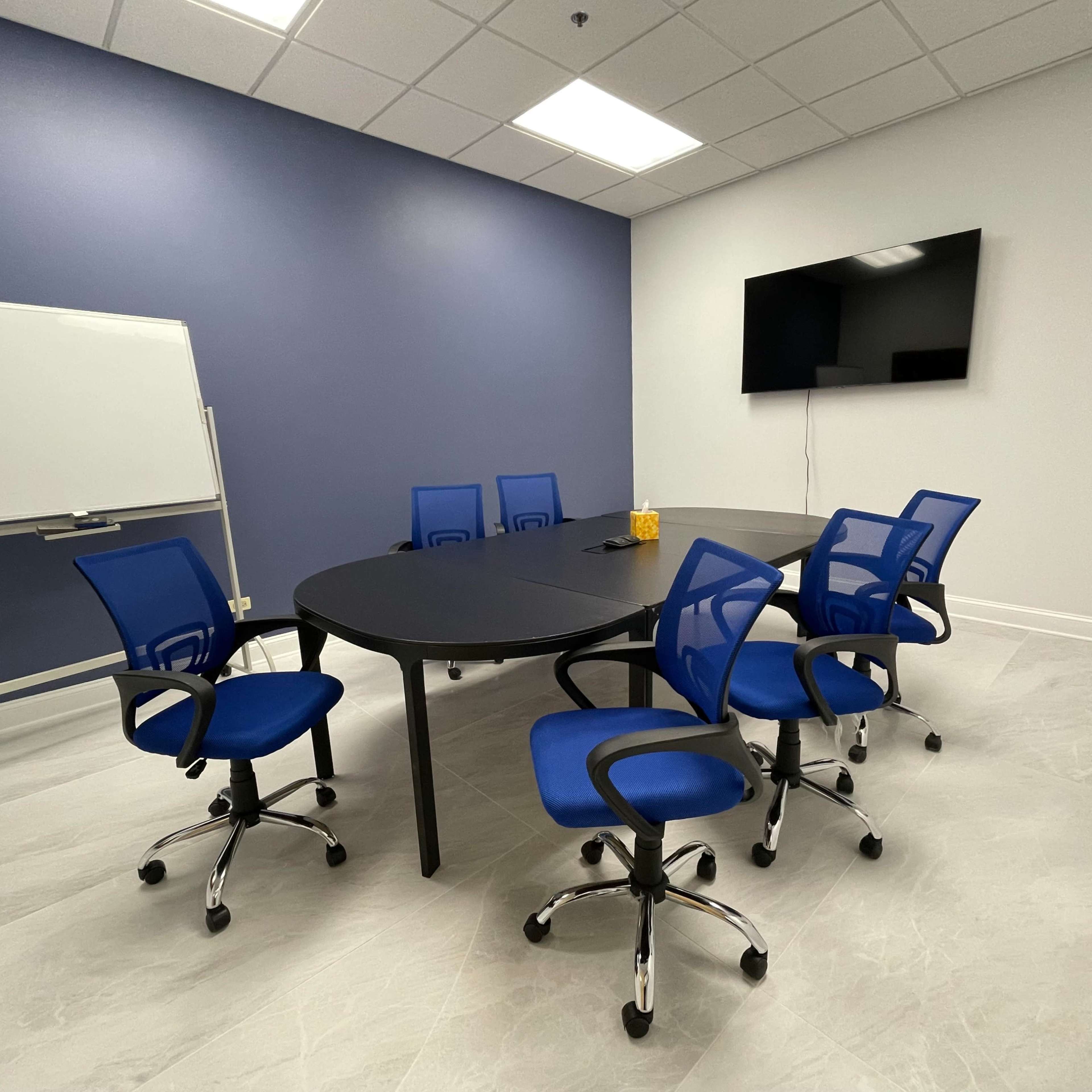 A modern conference room features a large oval table surrounded by six blue rolling chairs, a whiteboard, and a wall-mounted television.