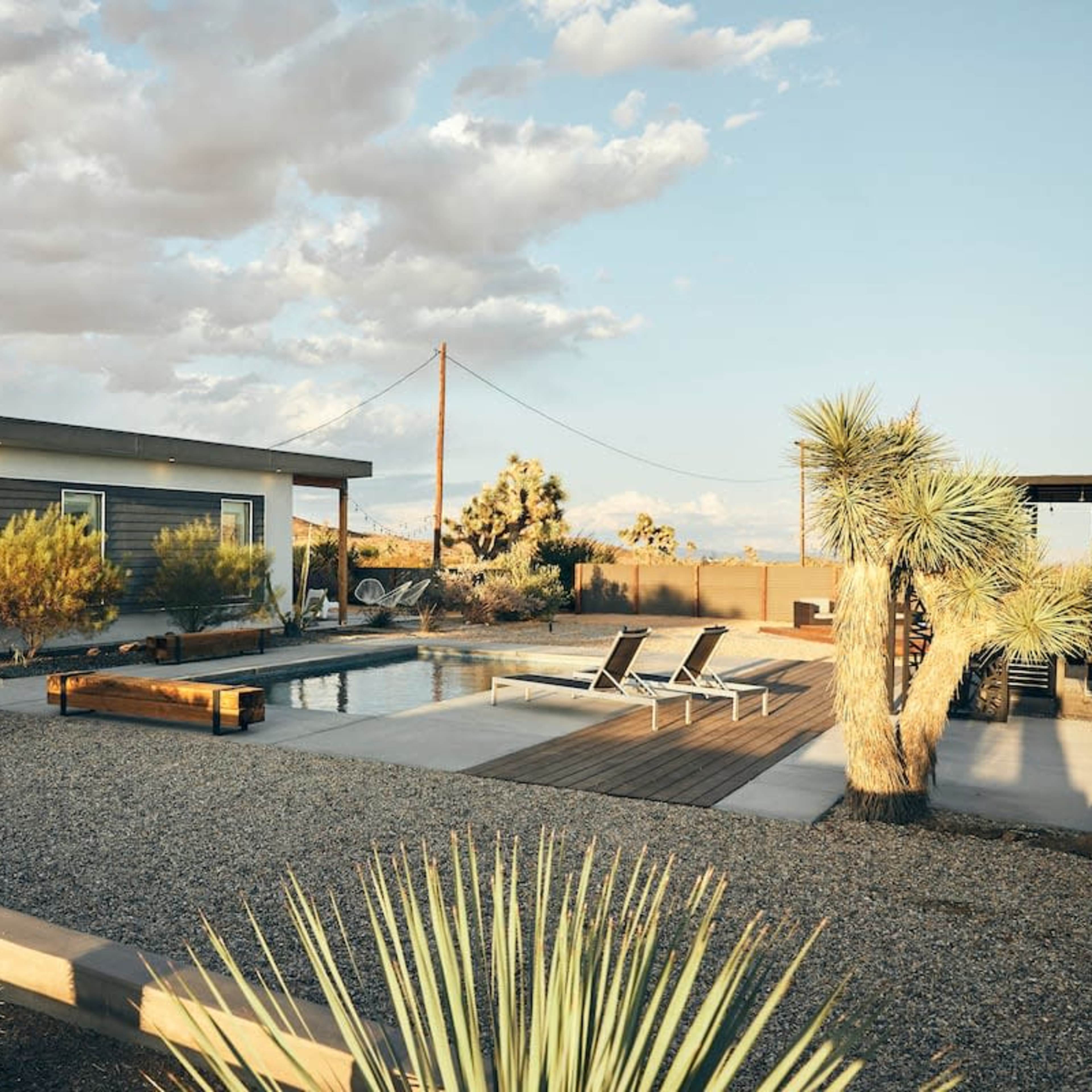 A modern home with a pool, surrounded by desert landscaping, features lounge chairs and various cacti under a partly cloudy sky.