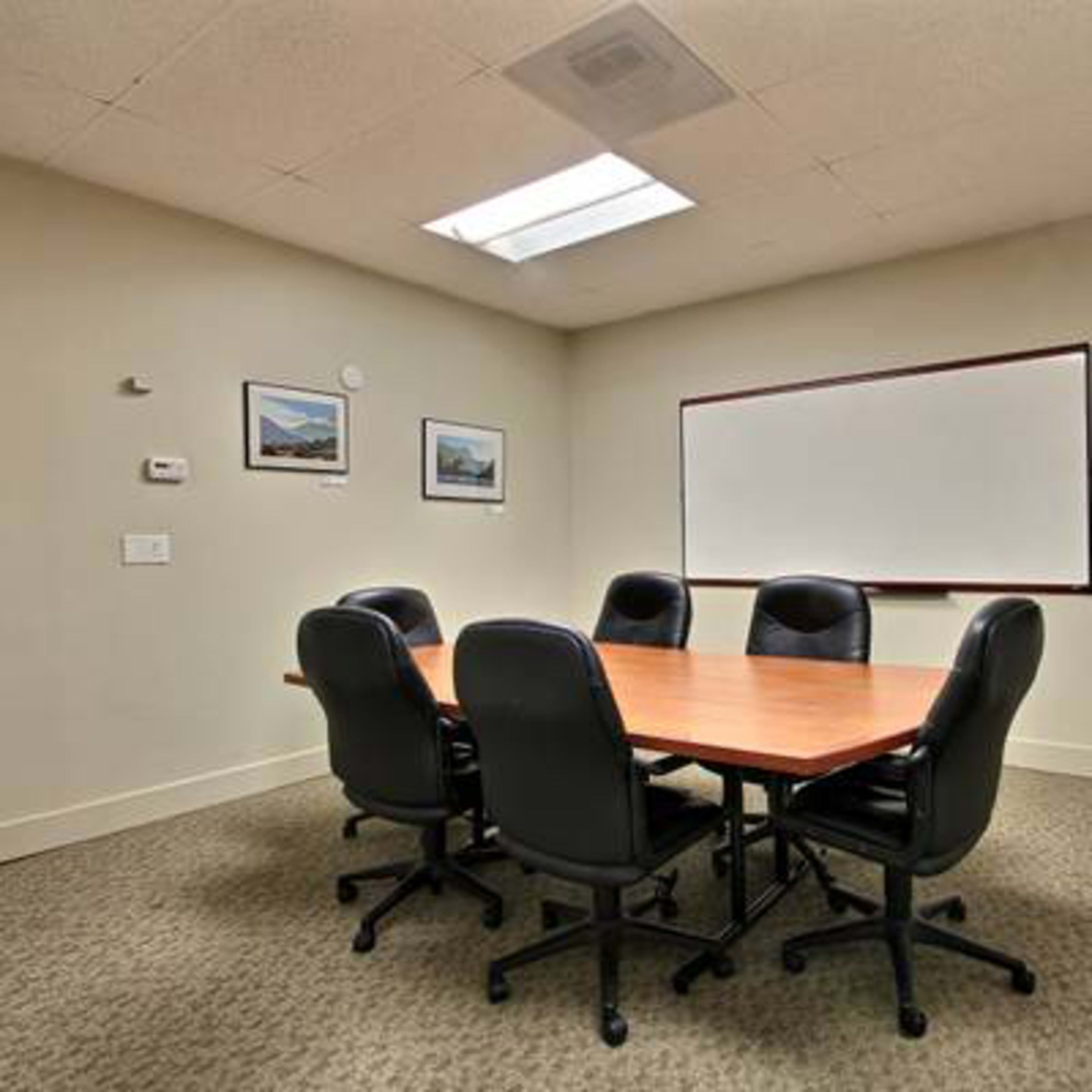 A small conference room features a rectangular wooden table surrounded by six black chairs, with a whiteboard and a television mounted on the wall.