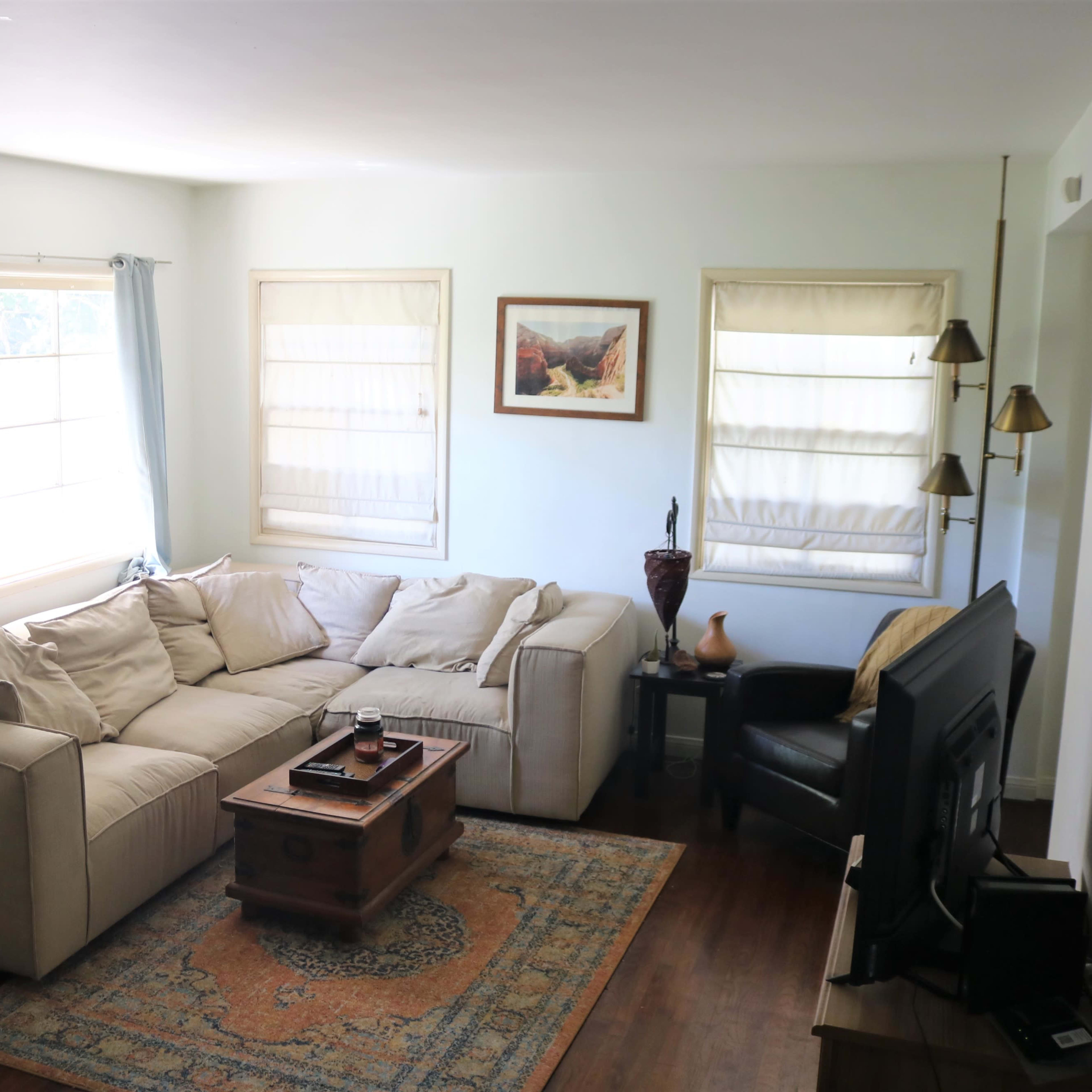 A cozy living room featuring a sectional sofa, a wooden coffee table, and a TV against the wall, with large windows allowing natural light to enter.