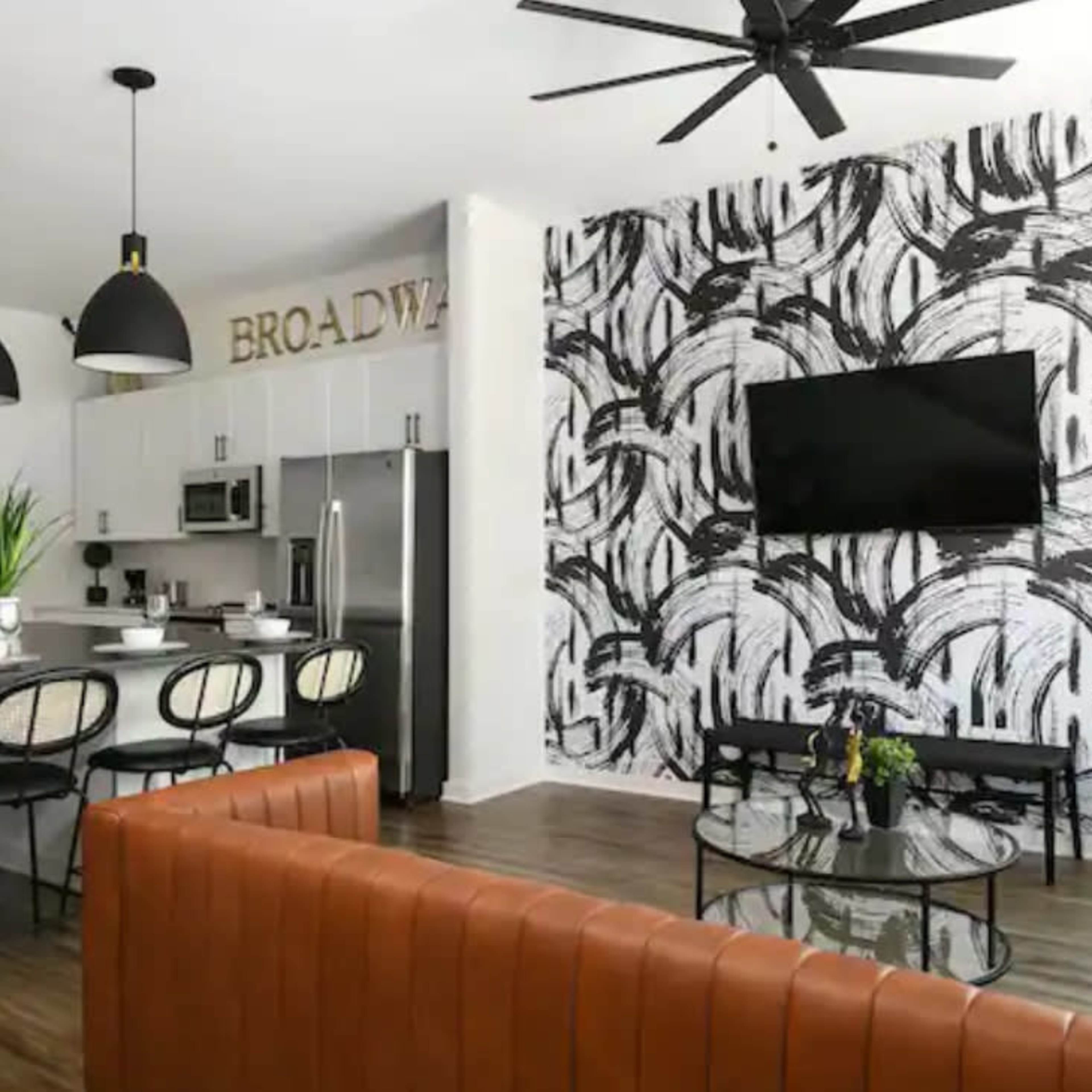A modern open-concept living area featuring a kitchen island with bar seating, a dining table set for several guests, and a lounge area with a brown leather sofa and a coffee table, all against a patterned accent wall and a flat-screen TV.