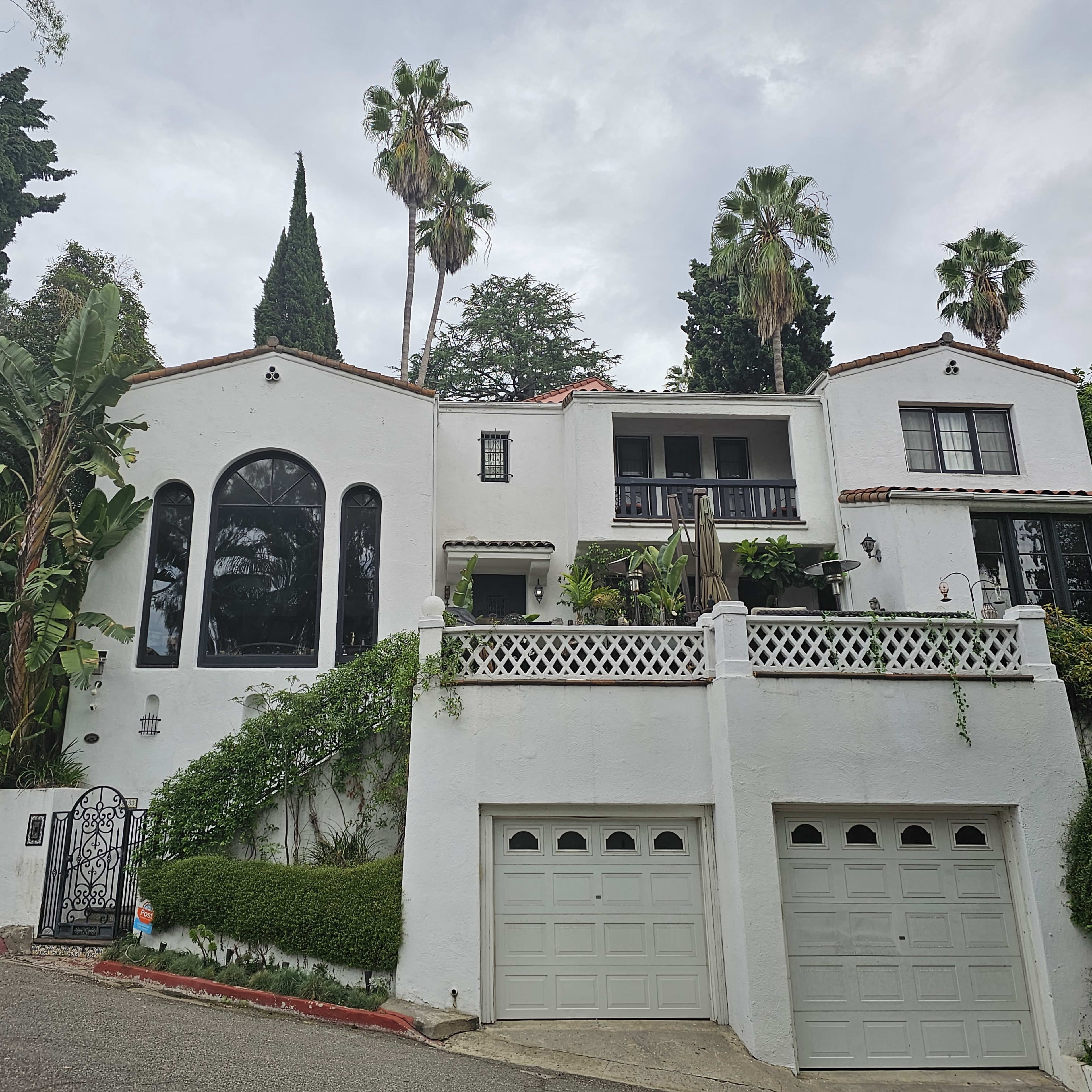 A white, two-story house with a prominent arched window and palm trees is situated on a sloped street.