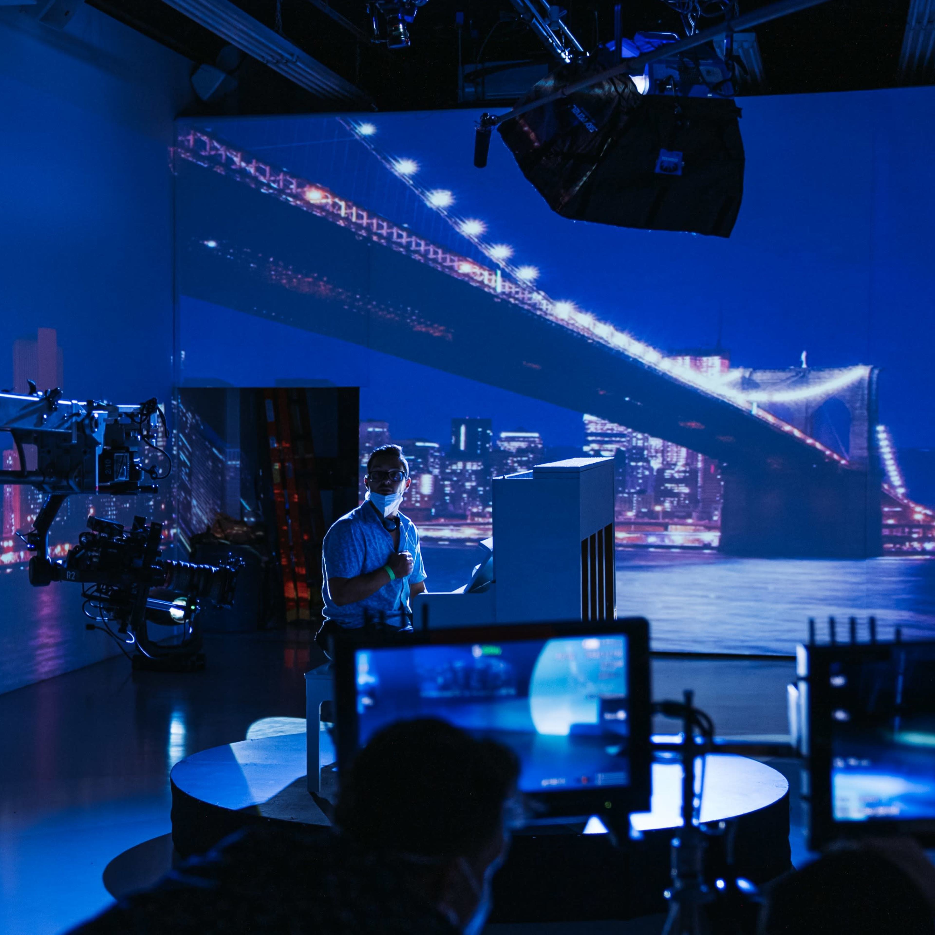 A television studio setup features a presenter on a circular podium with a backdrop of a brightly lit city skyline at night.