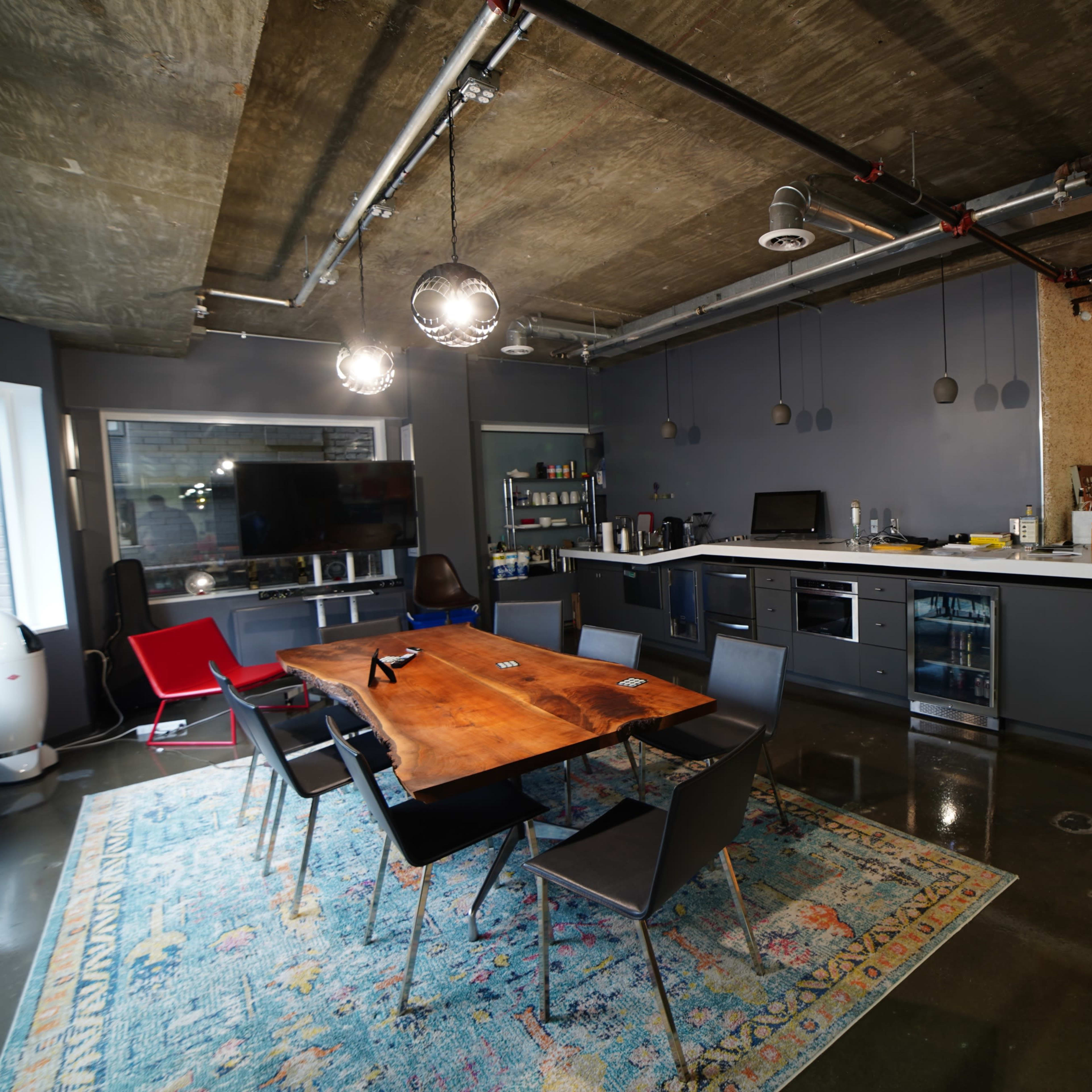 A modern, open-concept kitchen and dining area with a wooden table, black chairs, and sleek appliances, set against a backdrop of concrete walls and polished floors.